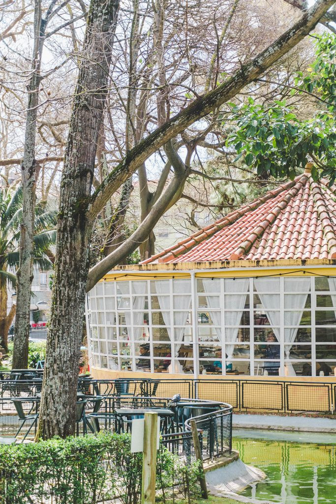 Outdoor café with white-curtained windows and tiled roof nestled in Lisbon’s Jardim da Estrela park, surrounded by tall trees and empty patio tables.