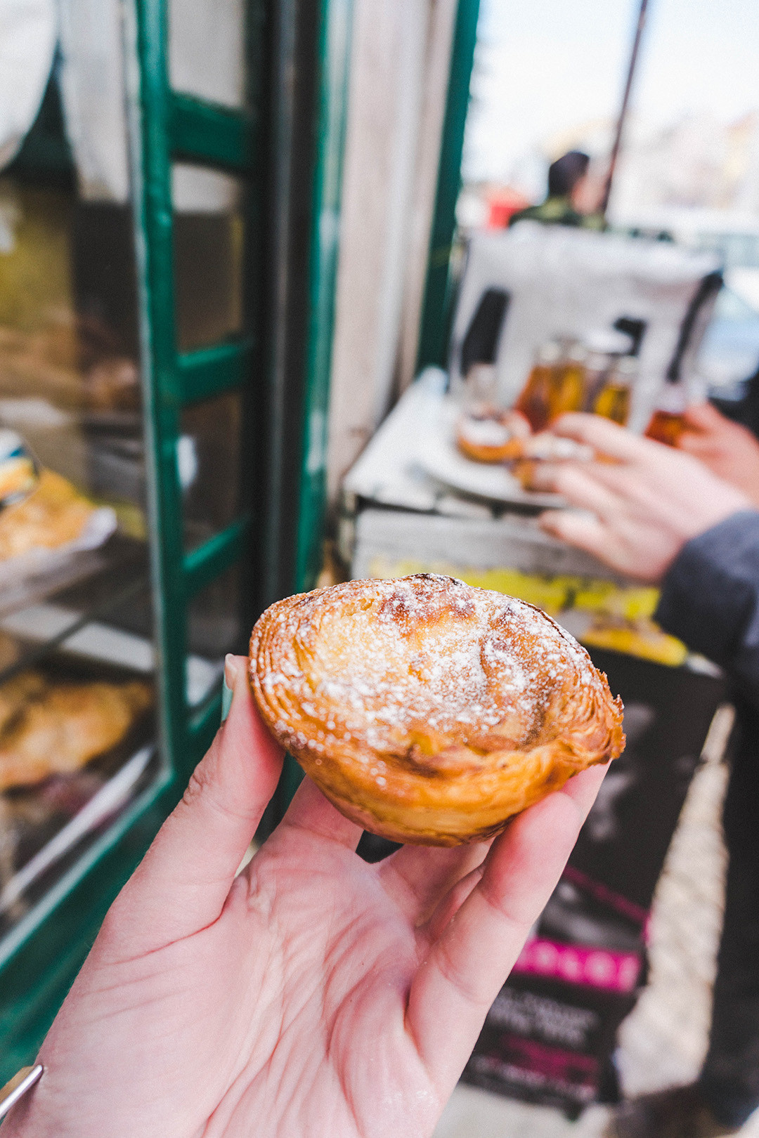 Close-up of a freshly baked pastel de nata dusted with powdered sugar, held in front of a local Lisbon bakery.