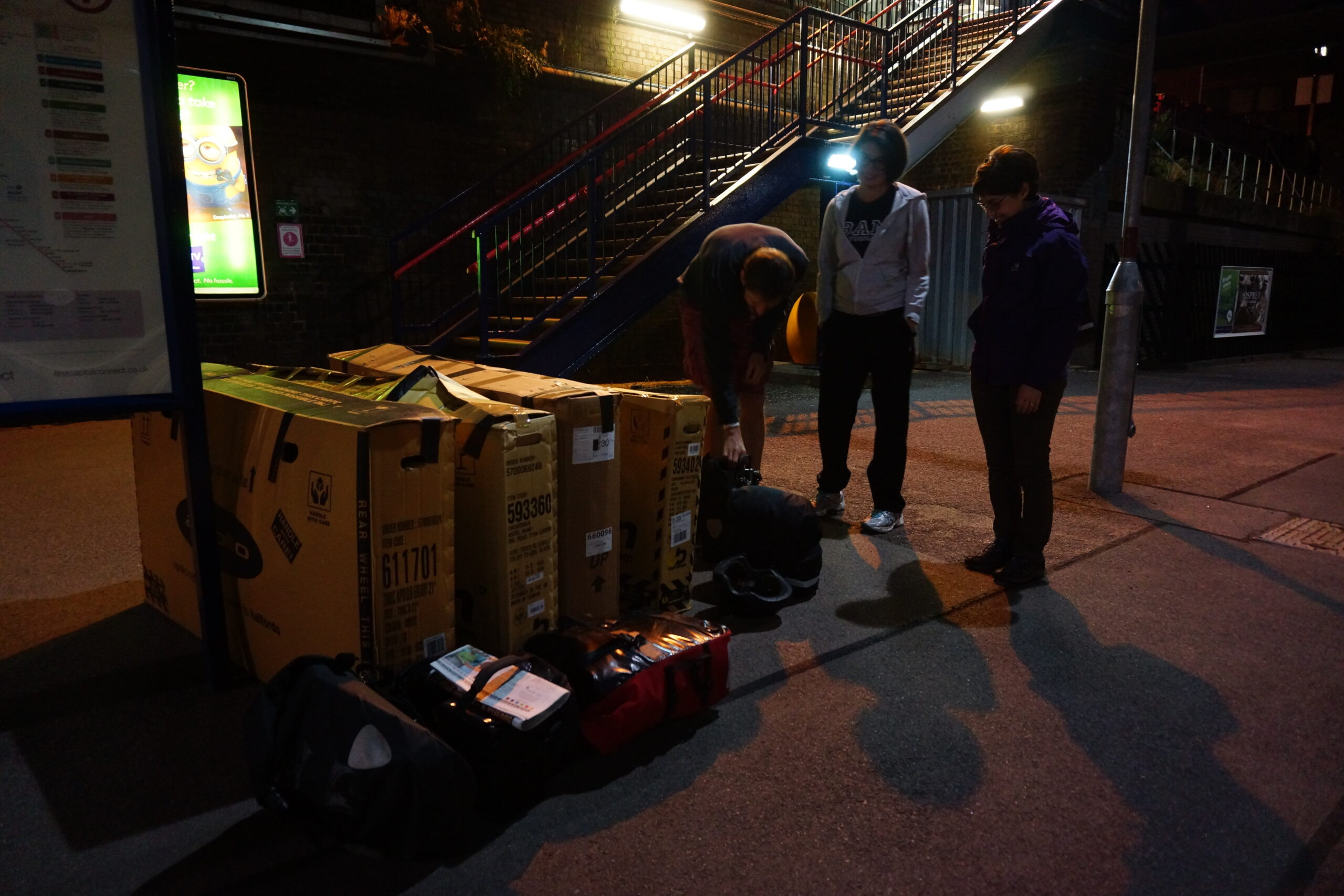 Four cyclists with boxed touring bikes waiting on a station platform in the dark.