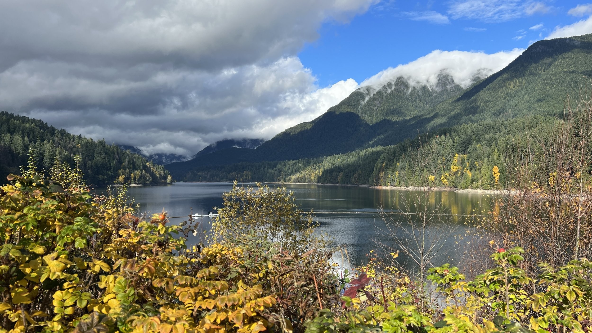 A calm lake surrounded by forested mountains under a partly cloudy sky, with autumn foliage in the foreground.
