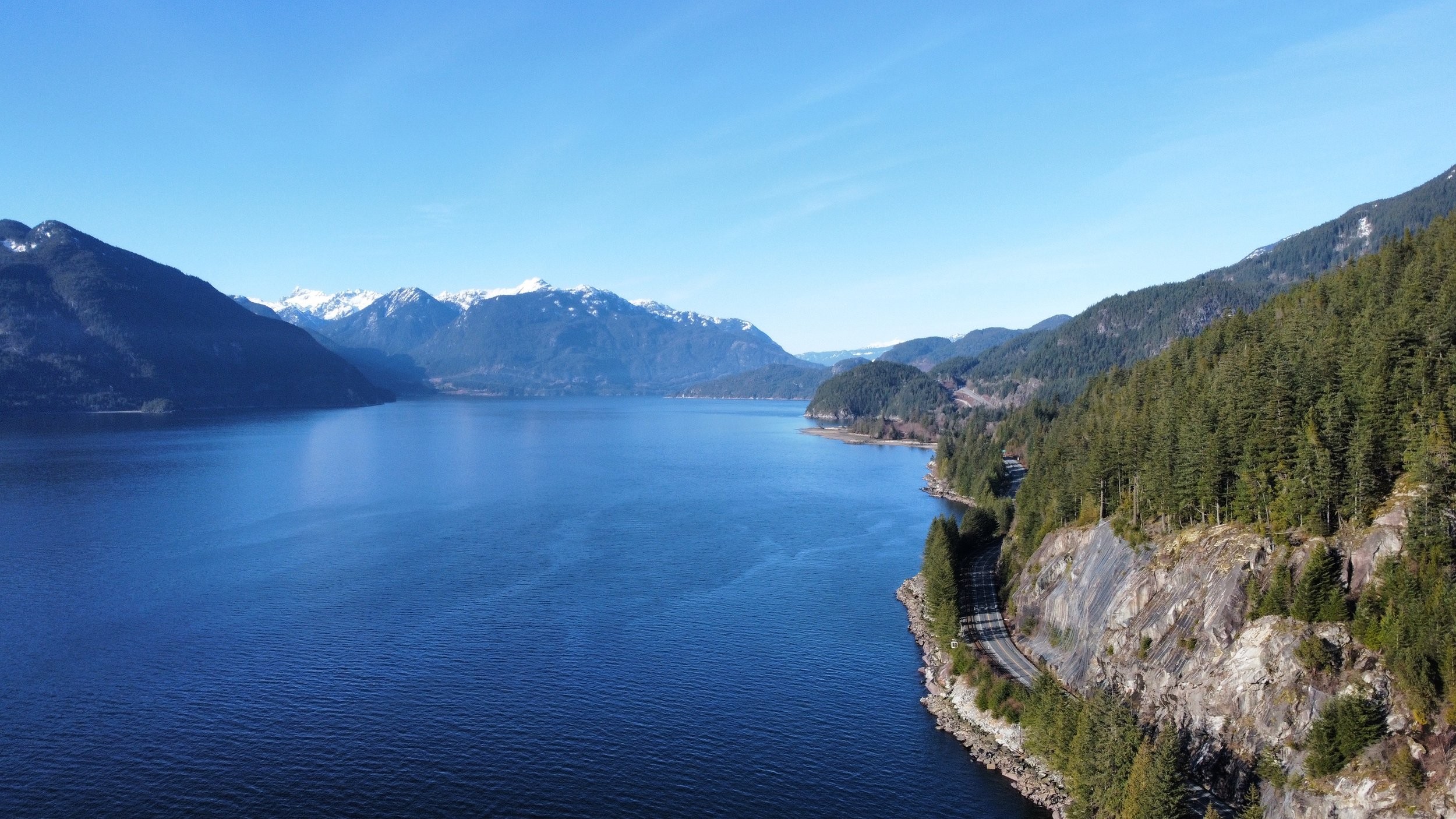 A wide river bordered by forested mountains and a road curving along the rocky shoreline under a clear blue sky.
