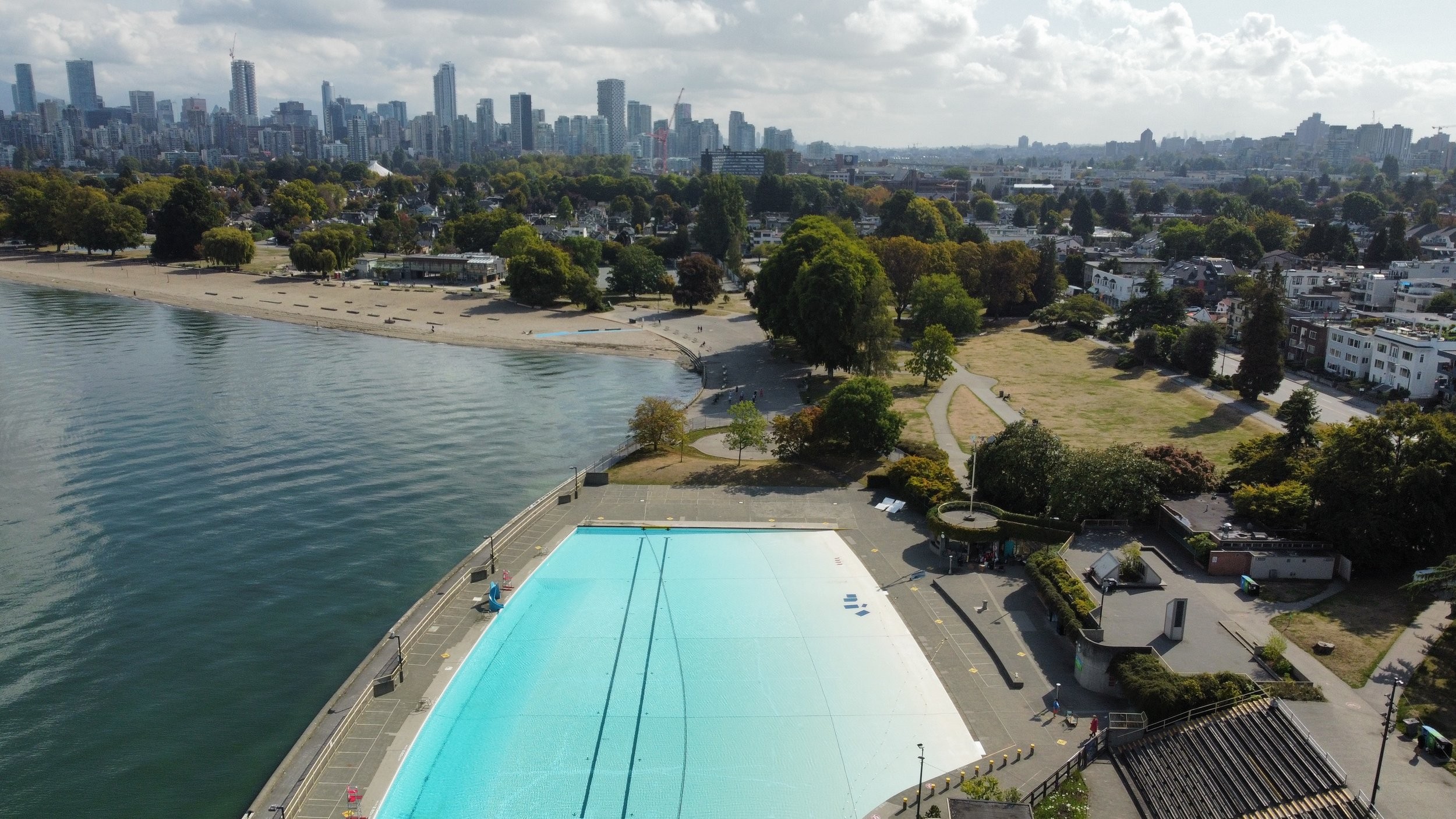 A large outdoor swimming pool by the waterfront with a city skyline and trees in the background under a partly cloudy sky.