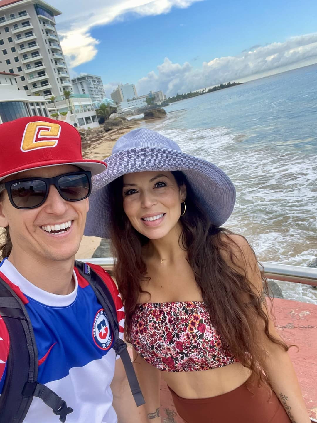 A couple taking a selfie on the beach, both smiling, with the ocean and sandy shore visible behind them.