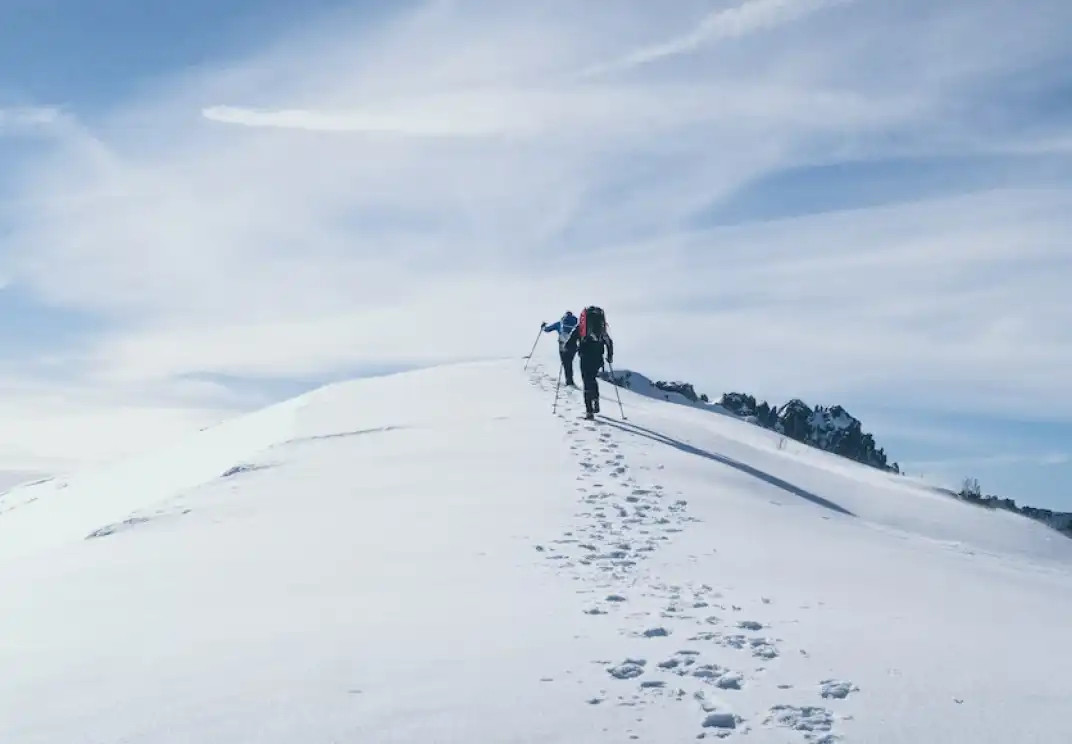 Two hikers climbing snowy mountain ridge with backpacks and trekking poles