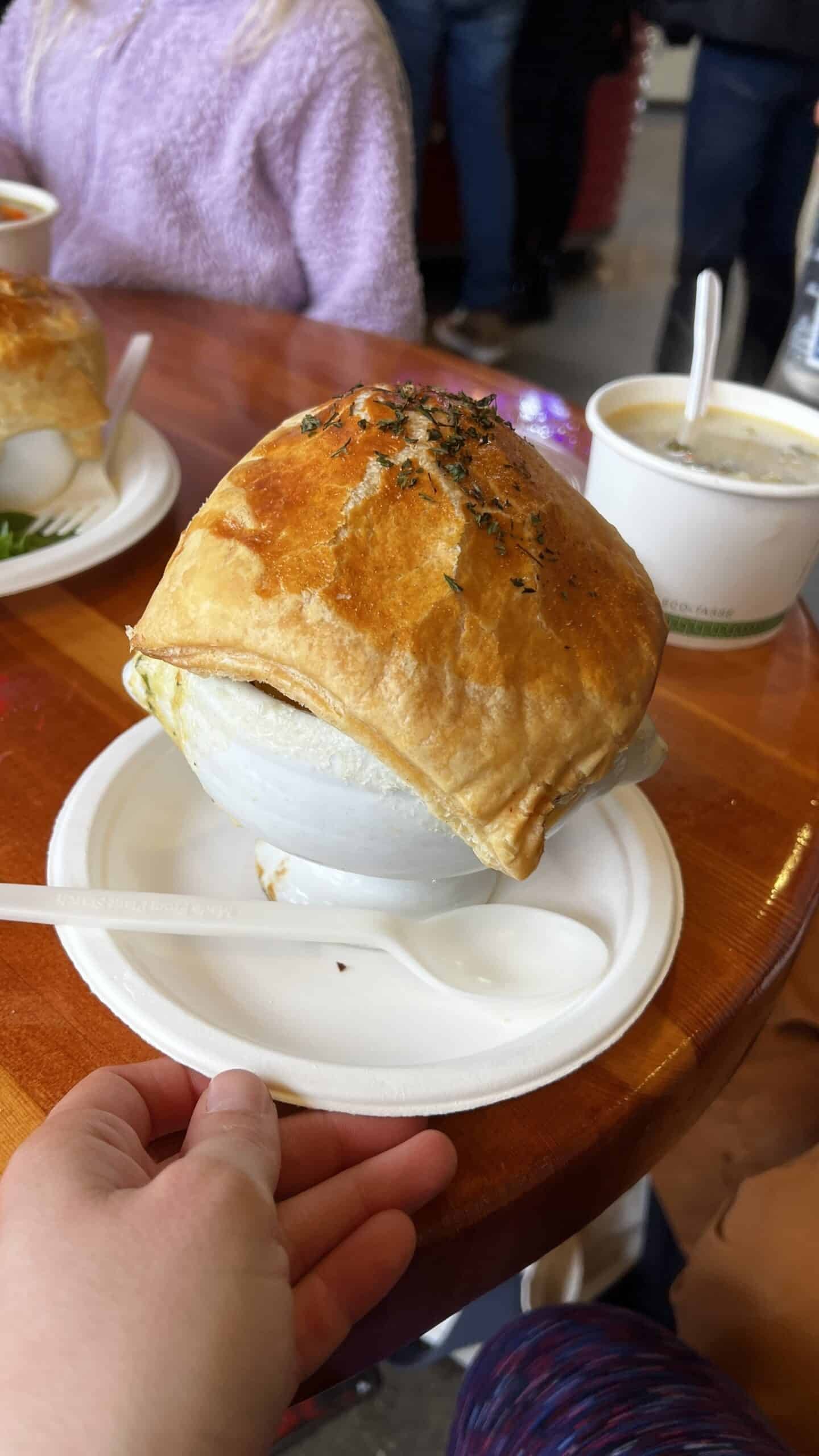 A bowl of soup with a puff pastry crust on top sits on a plate with a plastic spoon; a hand holds the plate.
