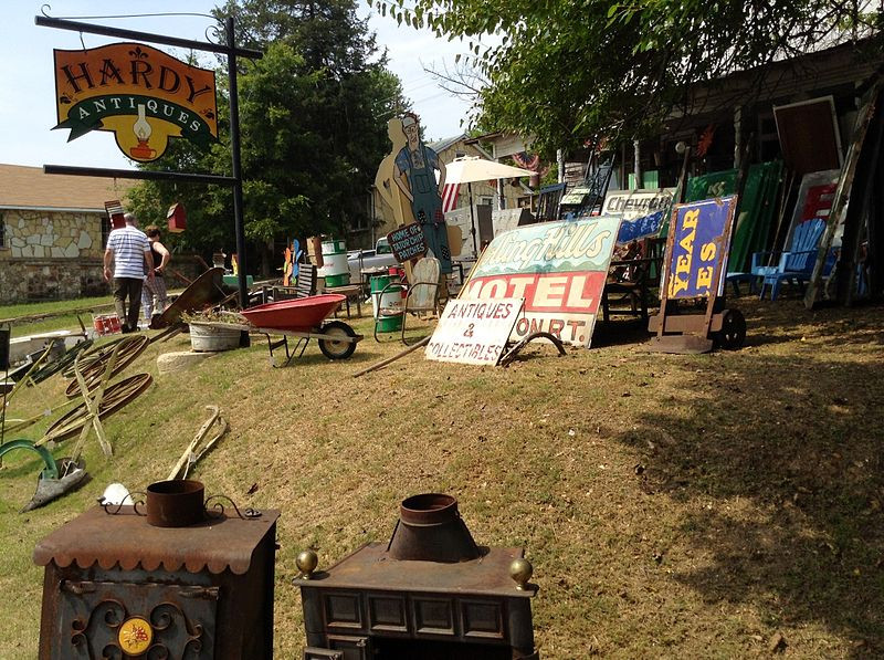 A vintage shop with various antiques, signs, and garden tools displayed on a grassy slope under a sunny sky.