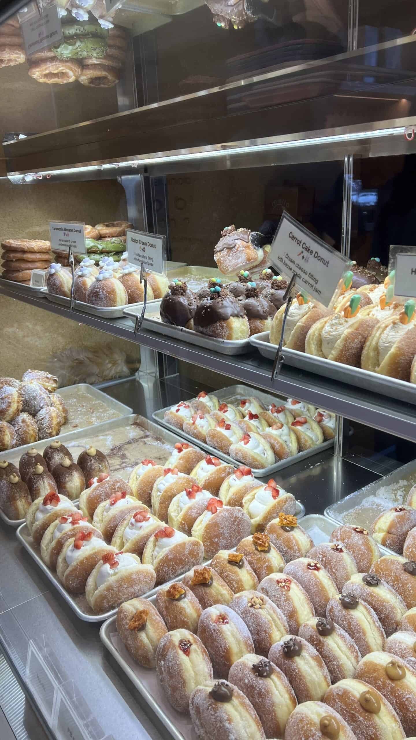 Display case filled with various filled donuts and pastries, some topped with fruit or chocolate, arranged in trays on multiple shelves.