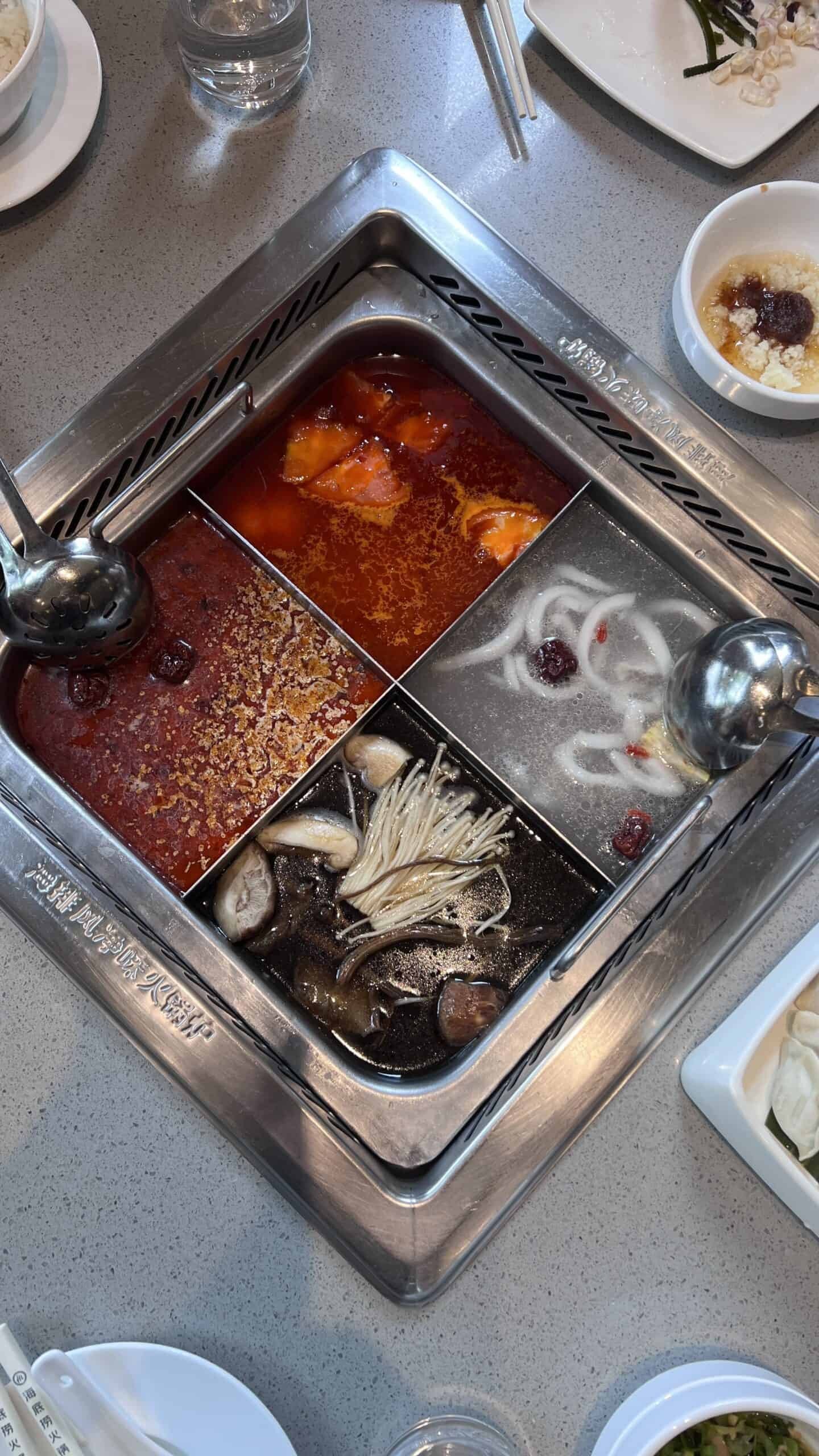 A metal hot pot divided into four sections with different broths and ingredients, surrounded by bowls and utensils on a dining table.