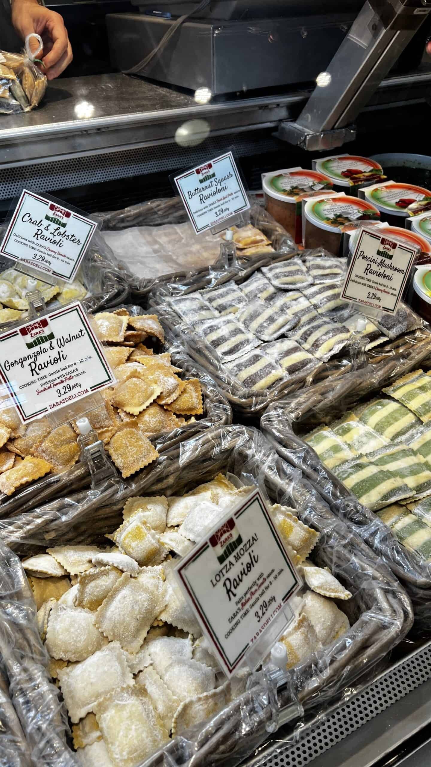 Assorted ravioli varieties displayed in trays at a deli counter, each labeled with ingredients and prices.