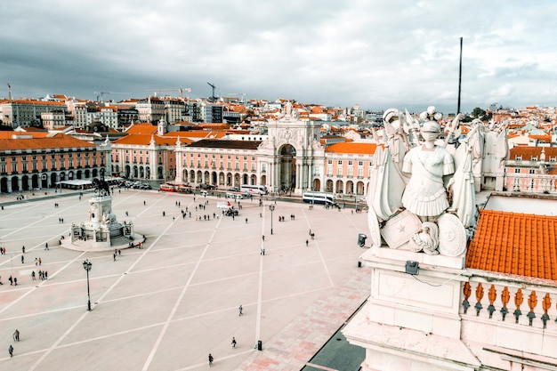 Large city square with statues, archway, and red-roofed buildings under a cloudy sky, people walking below.
