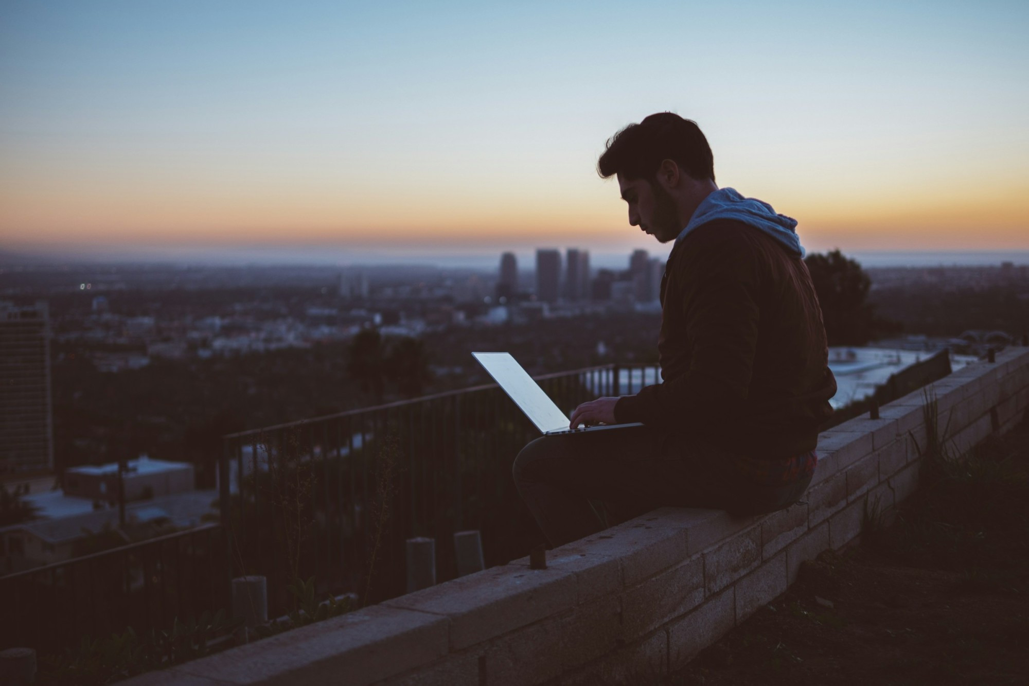 A man sitting on a rooftop at dusk, working on his laptop with an urban skyline in the distance