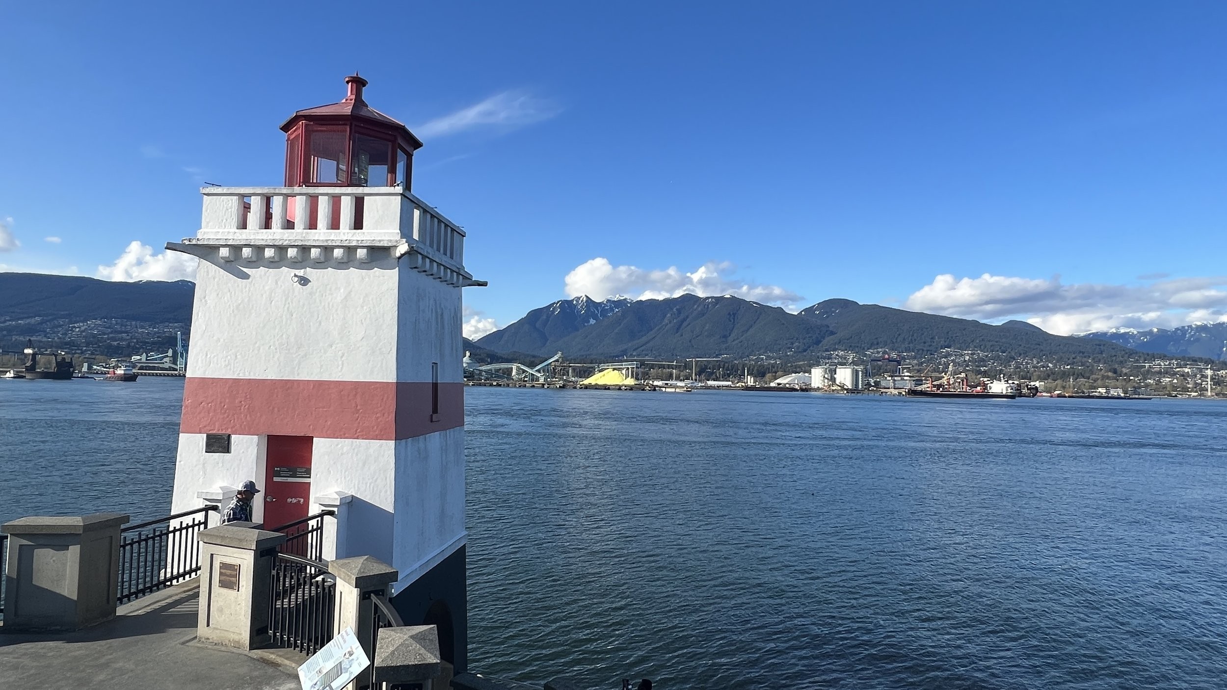 White and red lighthouse stands by the waterfront with mountains, industrial buildings, and clear blue sky in the background.