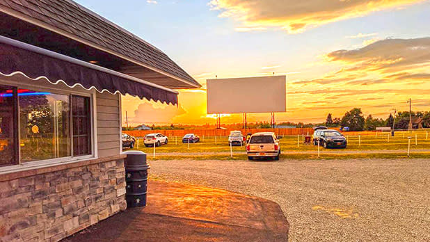 The exterir of a stone building with a giant outdoor screen in the distance and cars parked on the lawn at the Bay Drive in with the sun setting behind the screen