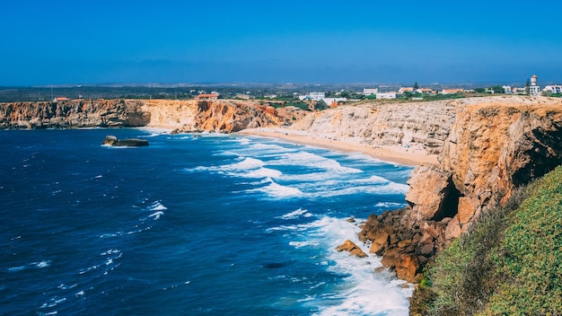 Rocky coastline with sandy beach, blue ocean waves, and distant buildings under a clear sky.