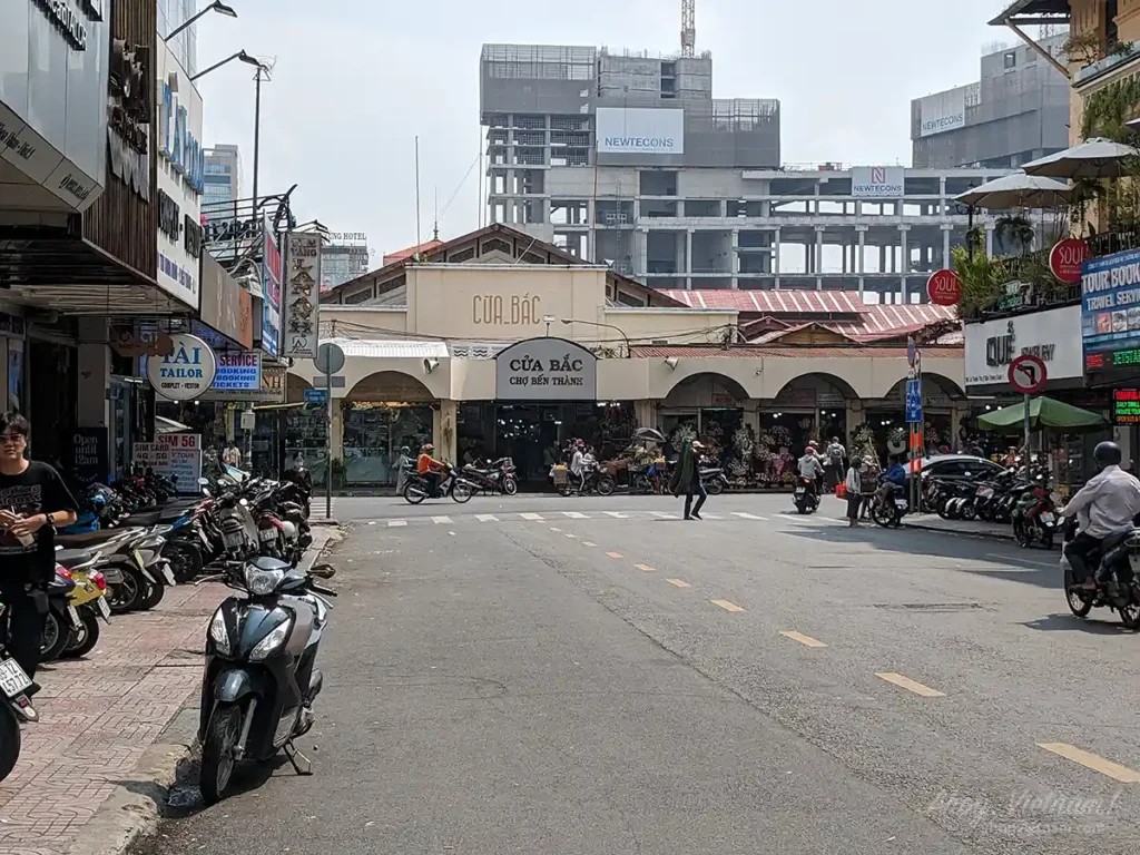 Thu Khoa Huan Road leading to Ben Thanh Market in District 1, Ho Chi Minh City. Motorbikes parked on side of street, shops, neon lights, and locals and tourists wandering around. 