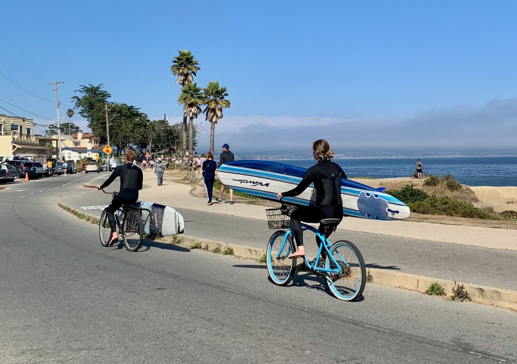 two people on bikes carrying surfboards. one person is carrying a big surfboard in her hand. The other person has a surfboard rack carrying the surfboard.