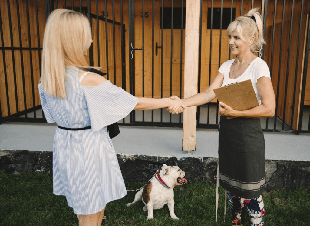 Two women shake hands at a dog boarding facility. One woman has a bulldog on a leash.
