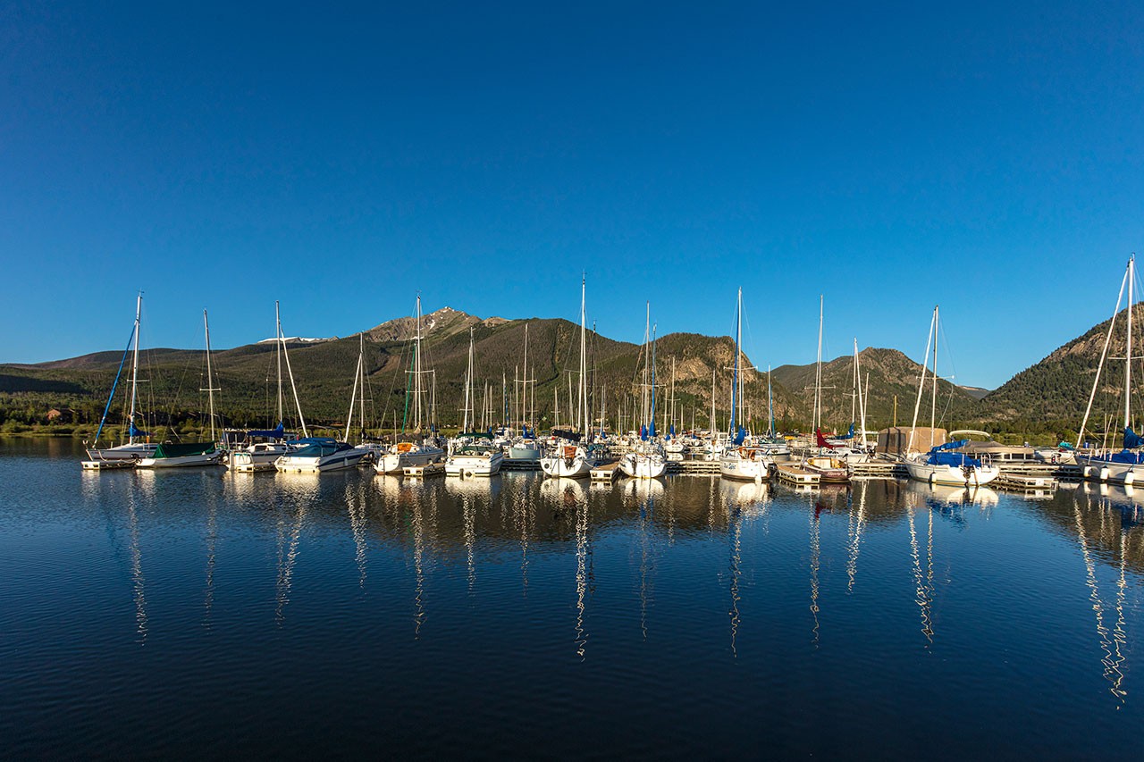 Docked sailboats with masts reflecting on dark blue lake, Peak One in the background.