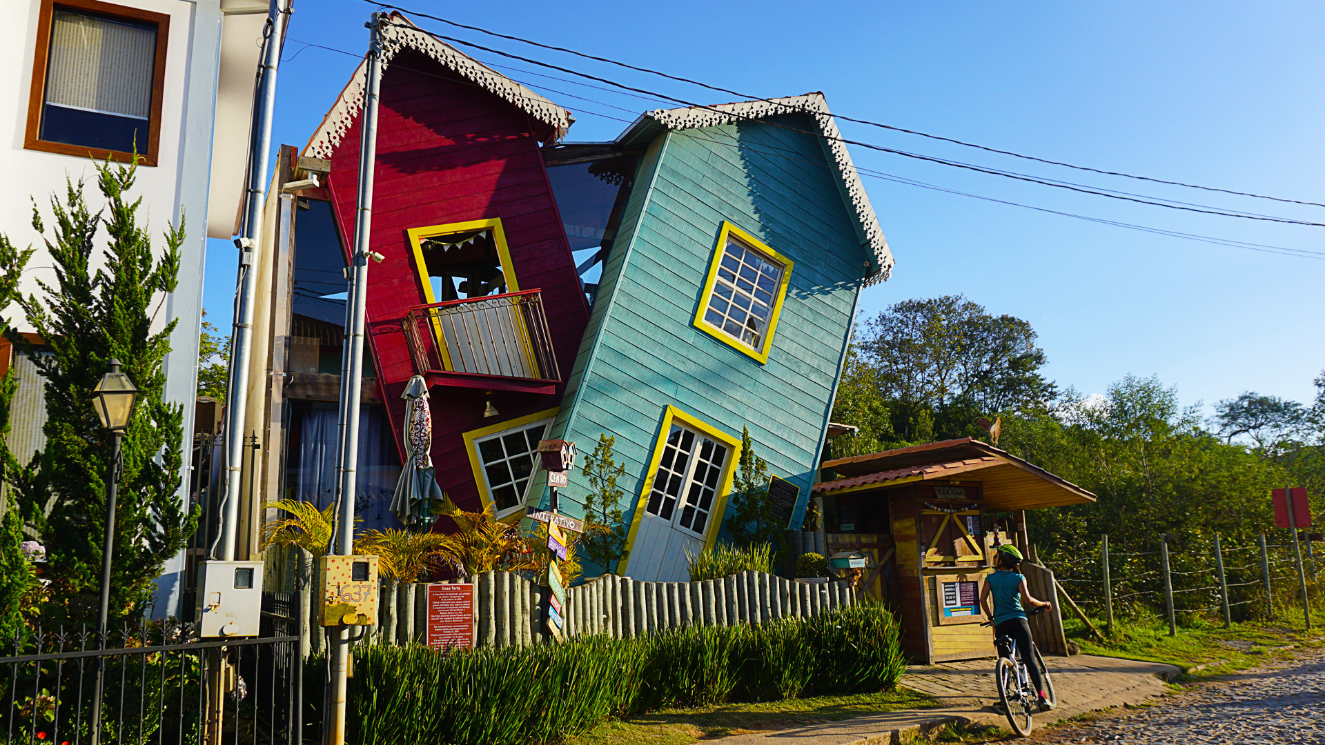 riding bicycles on cobble stone street with helmets on. There is a crooked house on one side. Bichinho, Tiradentes