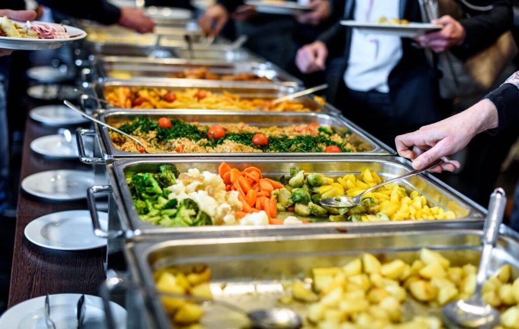 A variety of travel-friendly foods, including soup in a thermos, a casserole in an insulated container, a sandwich wrapped in parchment paper, and desserts packed in sturdy containers