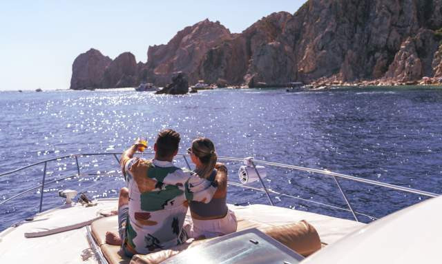 A couple relaxes on the deck of a yacht, embracing and pointing towards the scenic coastline of Los Cabos under a bright blue sky.