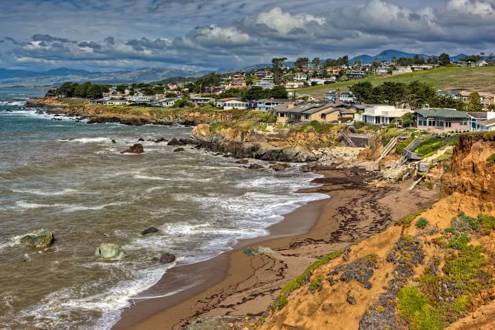A scenic coastal view of Cambria, California, featuring a sandy beach with gentle waves and a backdrop of rolling hills