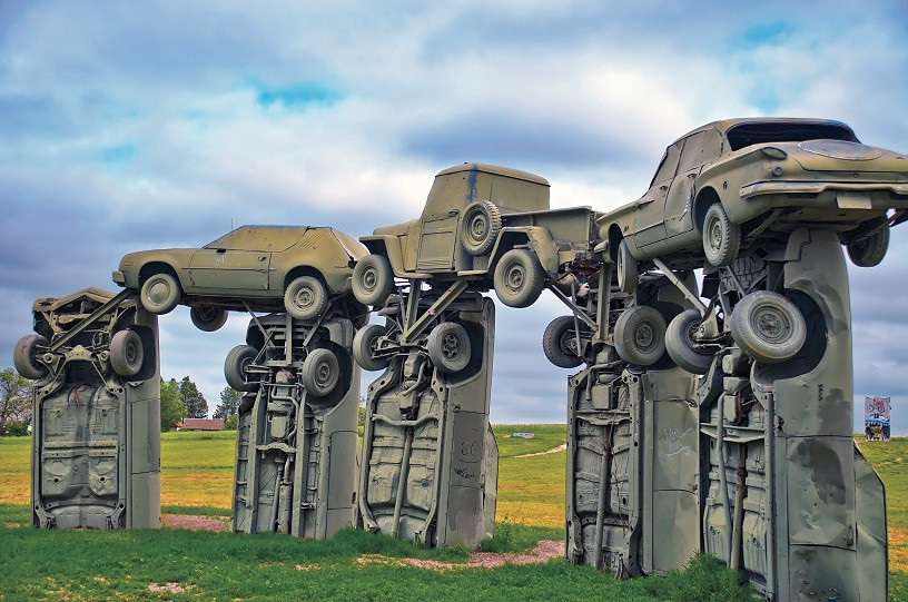 Carhenge Alt Text: Carhenge, an artistic replica of Stonehenge in Nebraska, constructed with vintage automobiles painted gray, standing in a field under a bright sky, drawing tourists interested in quirky roadside attractions.