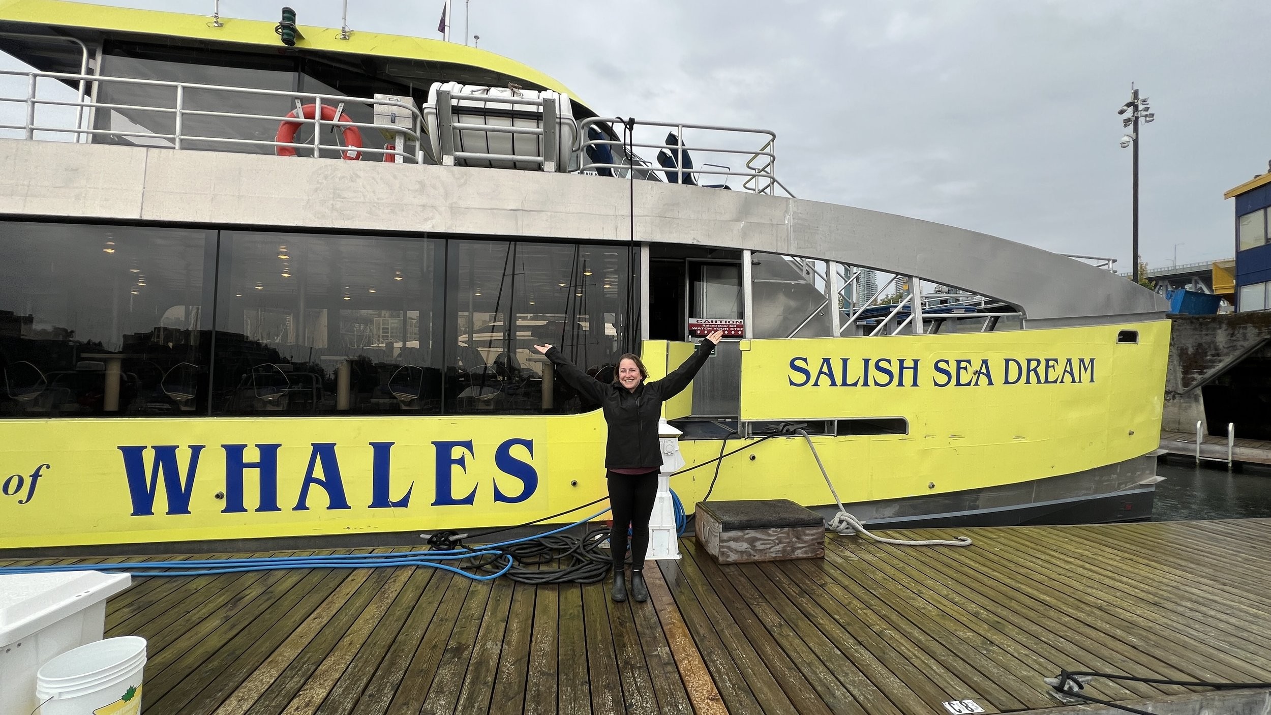 A person stands on a dock with arms raised in front of a yellow whale-watching boat labeled SALISH SEA DREAM.