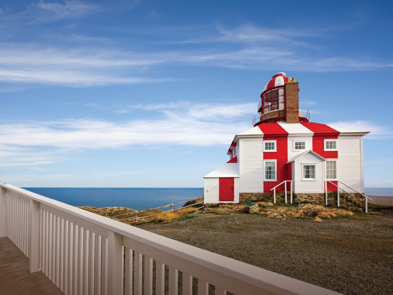 A white and red lighthouse stands on a rocky coastline under a clear blue sky. The building has a unique tiered structure with a lantern room at the top. A white fence runs along the foreground, with the ocean in the background.