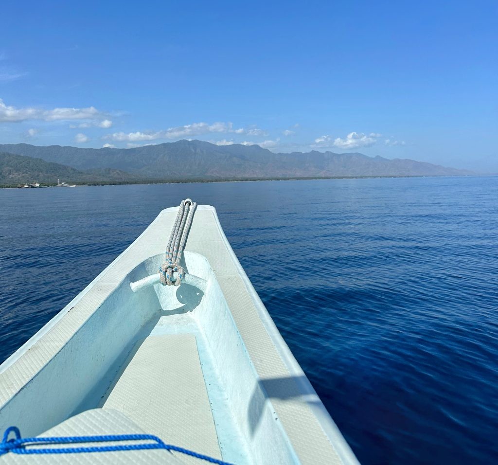A boat gliding through crystal-clear waters towards the coast of Bali, offering dolphin tours with a backdrop of majestic mountains.