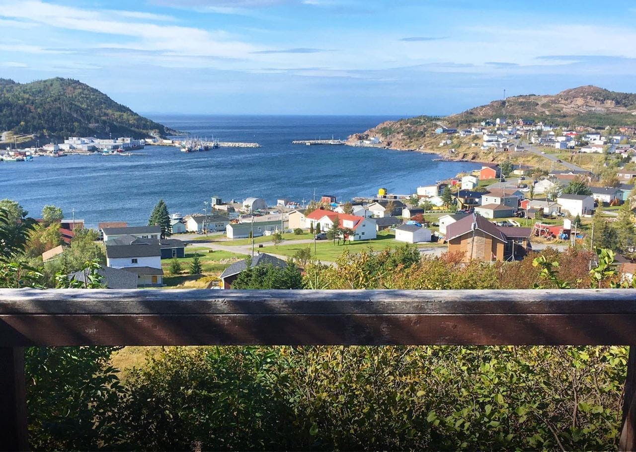 Scenic view from a wooden railing overlooking a coastal town with colorful houses. The town is nestled between two hills and borders a calm blue ocean under a clear sky. Vegetation in the foreground enhances the vibrant landscape.