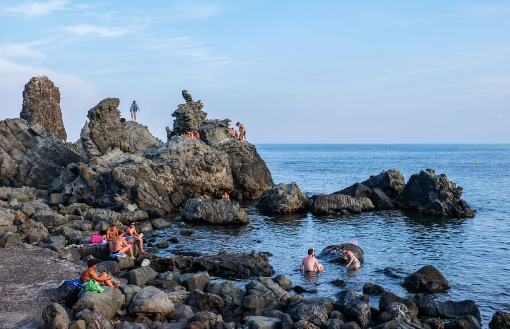 People hanging out on the rocky coastline in the blue water of Aci Trezza, Sicily
