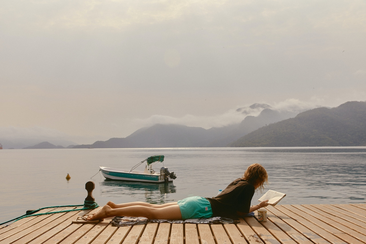 Woman in white long-sleeved shirt sitting on gray pavement near body of water during daytime