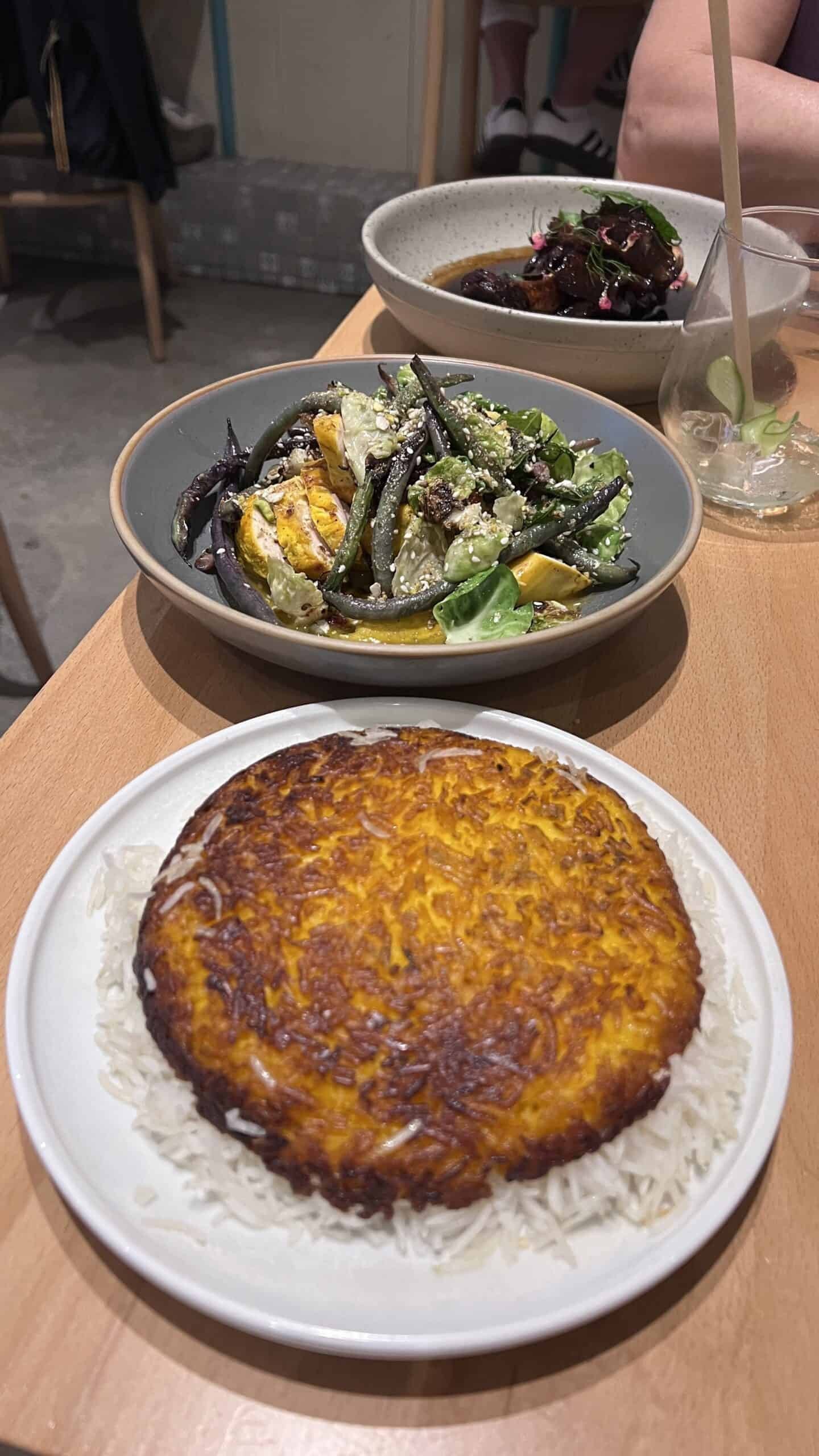 Three plates of food on a wooden table, featuring crispy rice, a vegetable dish, and a bowl of stewed food with herbs.