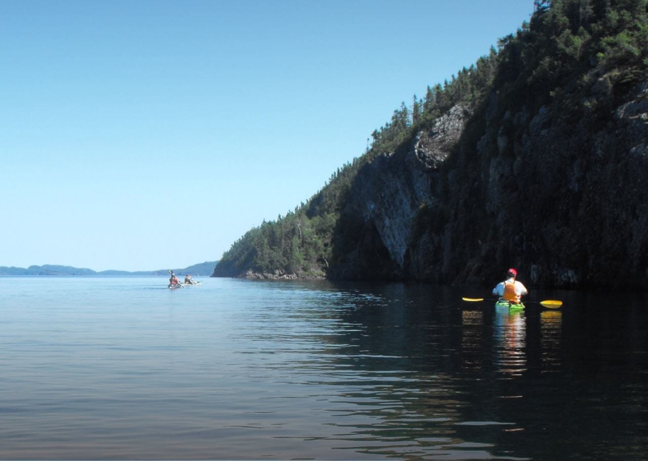 Two kayakers paddle through calm waters alongside a steep, tree-covered cliff. The sky is clear and blue, and distant islands are visible on the horizon.