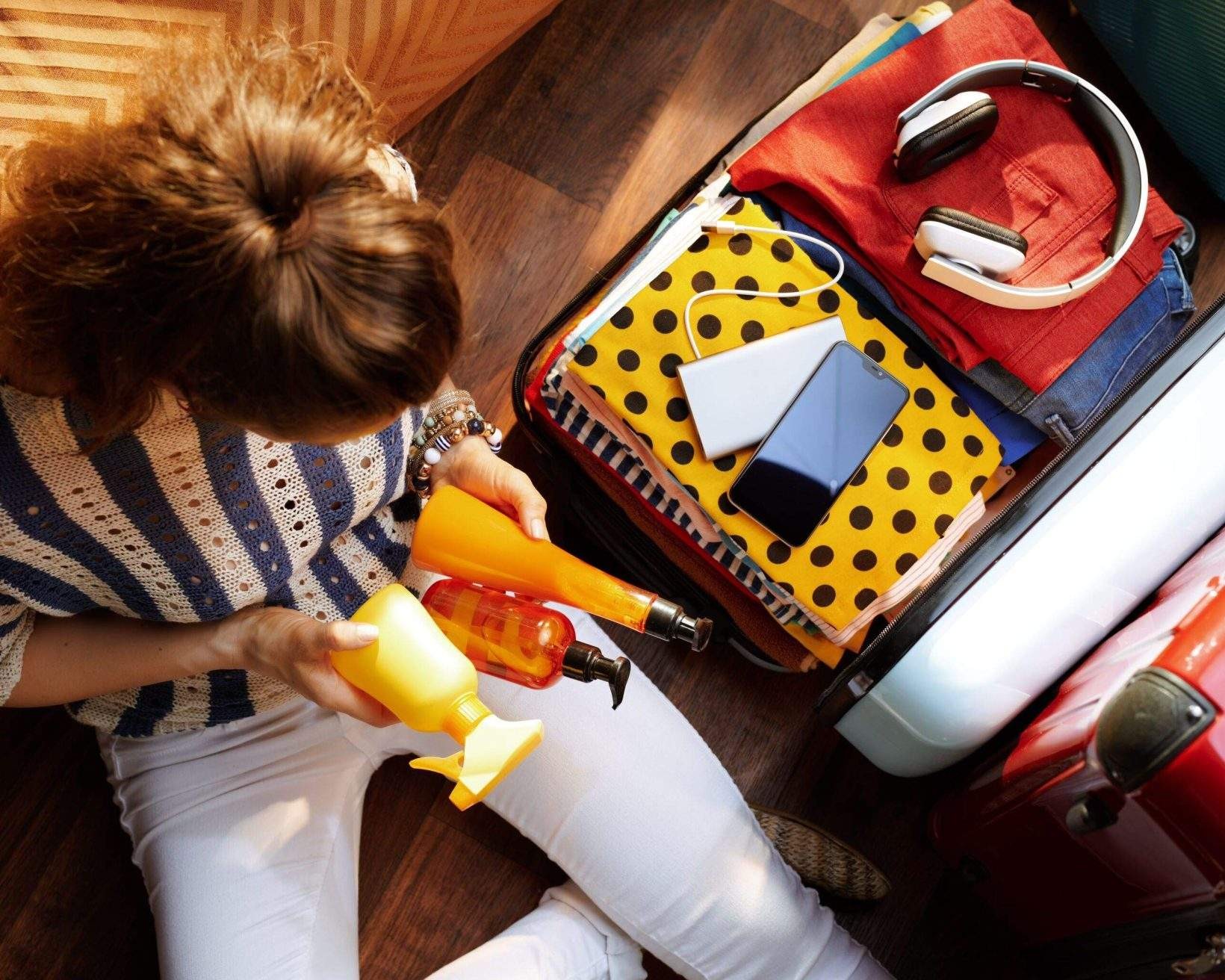 Upper view of modern woman in white pants and striped blouse in the modern house in sunny summer day packing summer cosmetics in open travel suitcase.