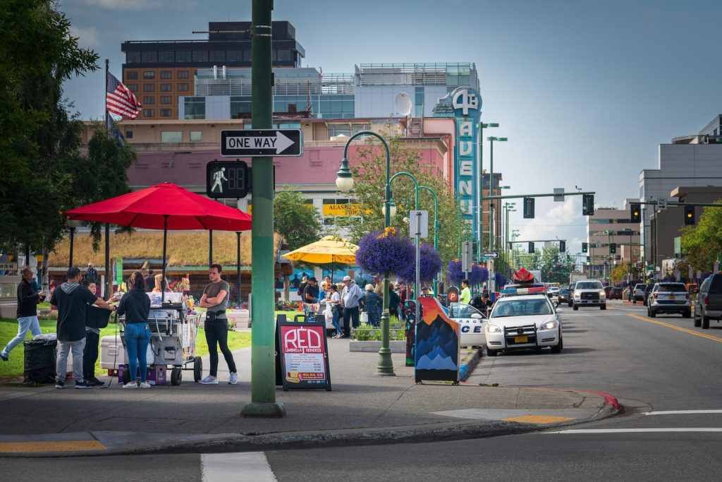 Vibrant street scene with pedestrians, vendors, and cars on a sunny day, featuring colorful umbrellas and a clear signpost indicating Fourth Avenue in Anchorage, Alaska
