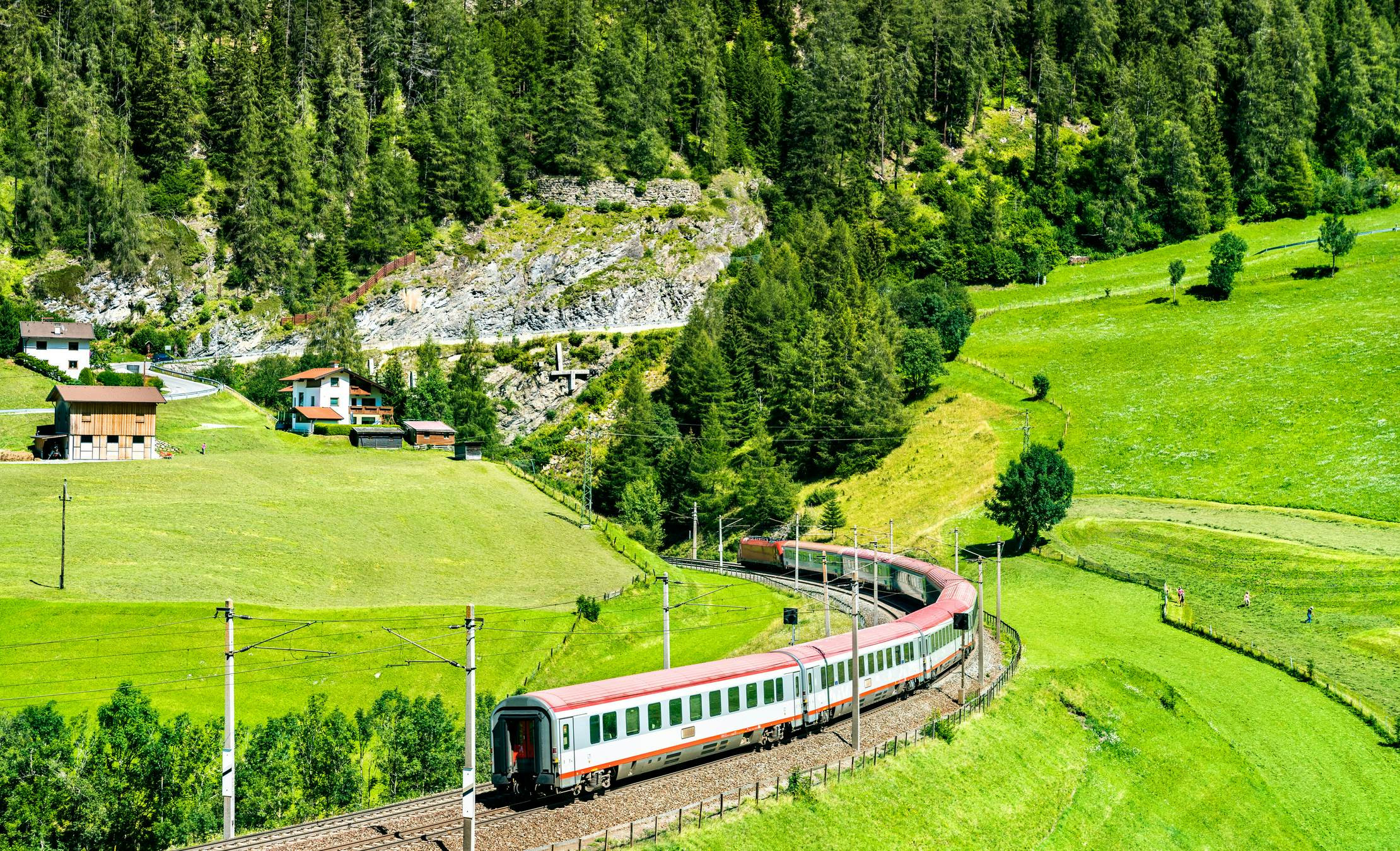 Train curving around a track through bright green fields with houses and forest in the background on a sunny day.