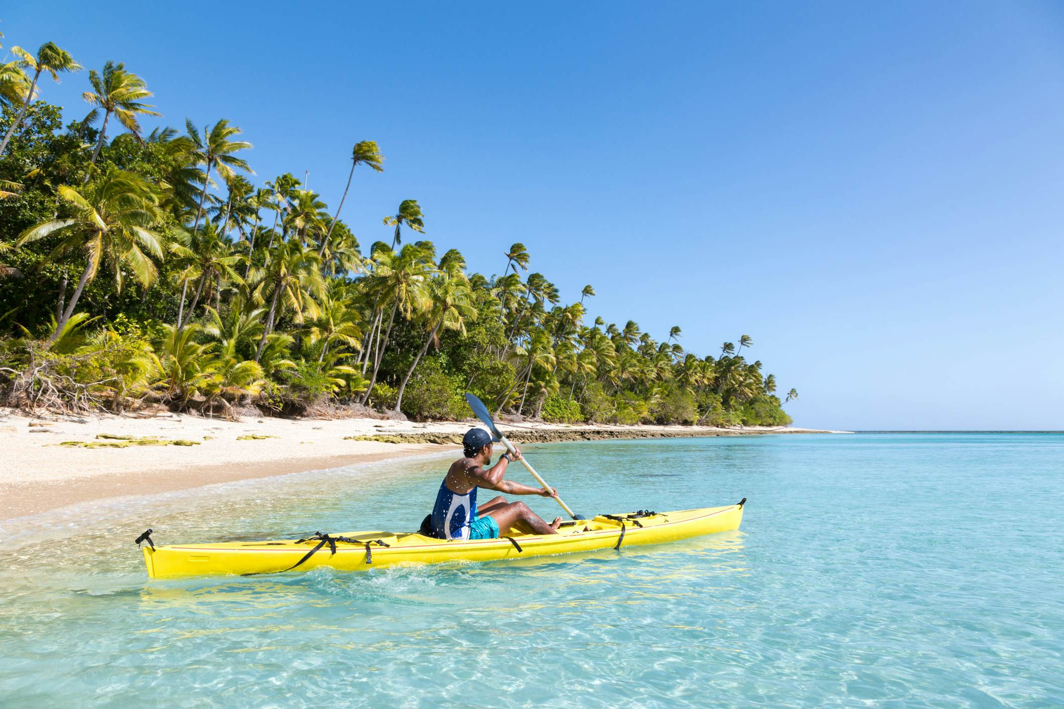 A man in a yellow kayak pushes off from a beach into the clear waters of the tropical sea.
