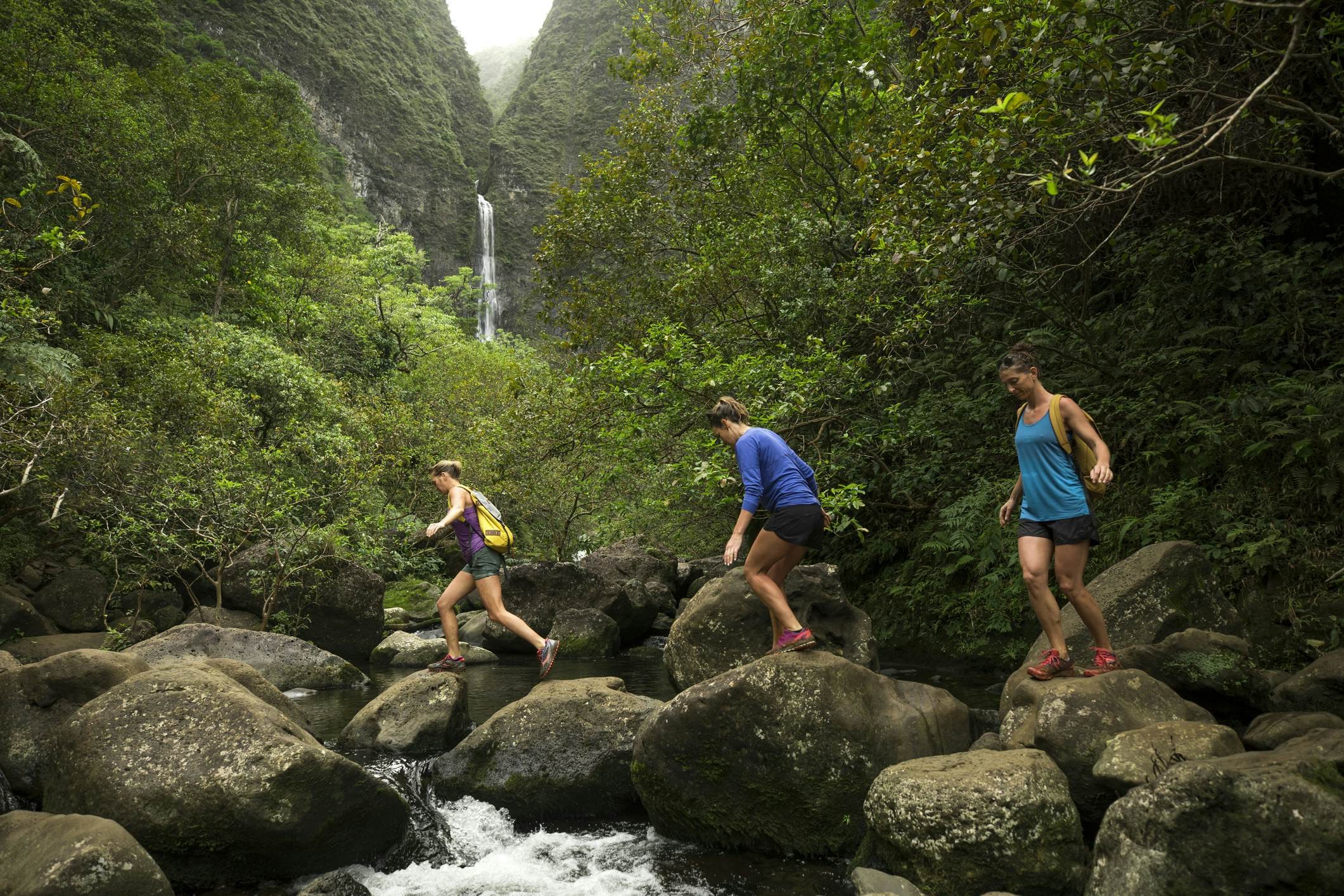 Three woman crossing water over large rocks in Hawaii, with a waterfall in the distance
