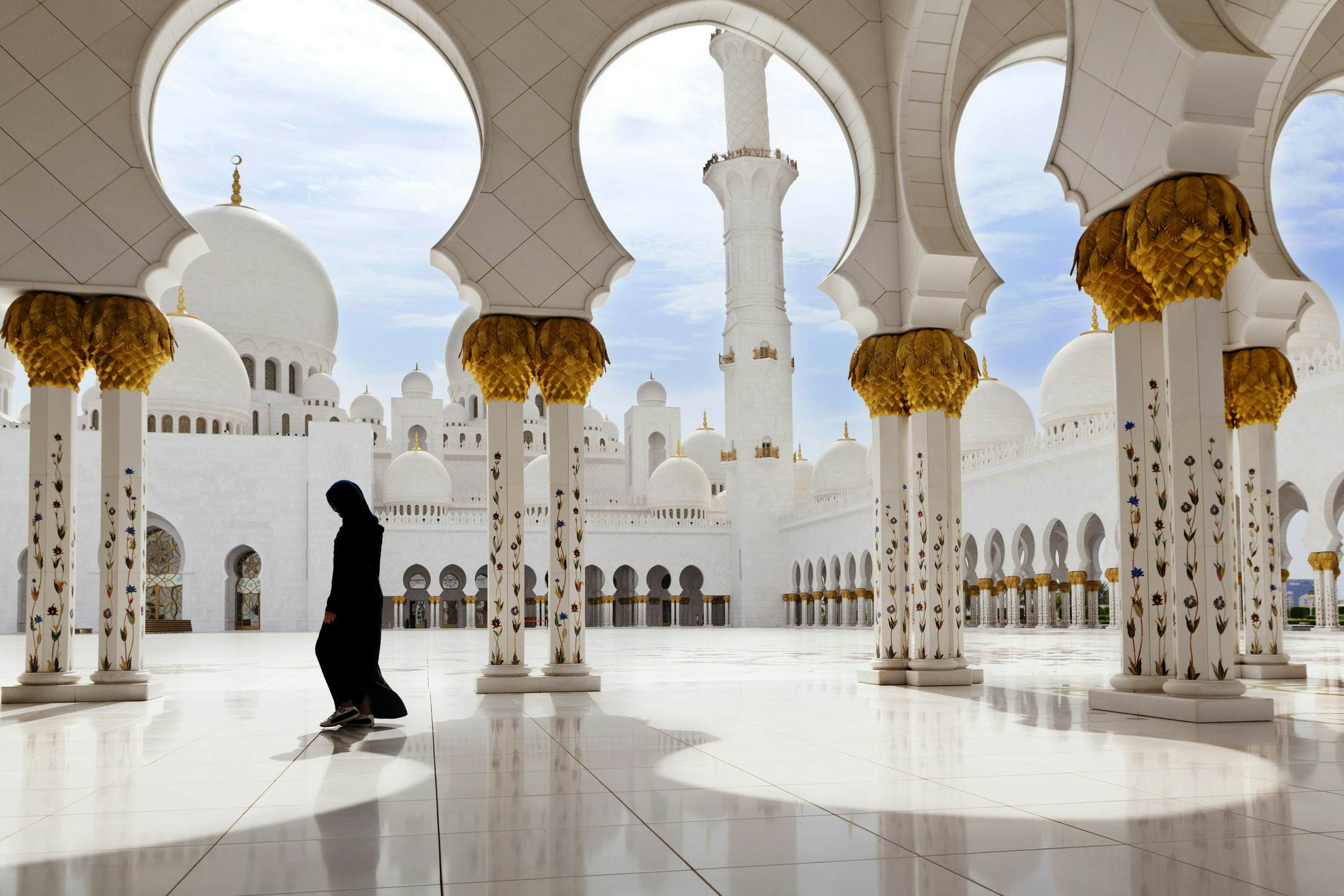 A woman wearing modest clothing stands in a courtyard of a vast white mosque complex