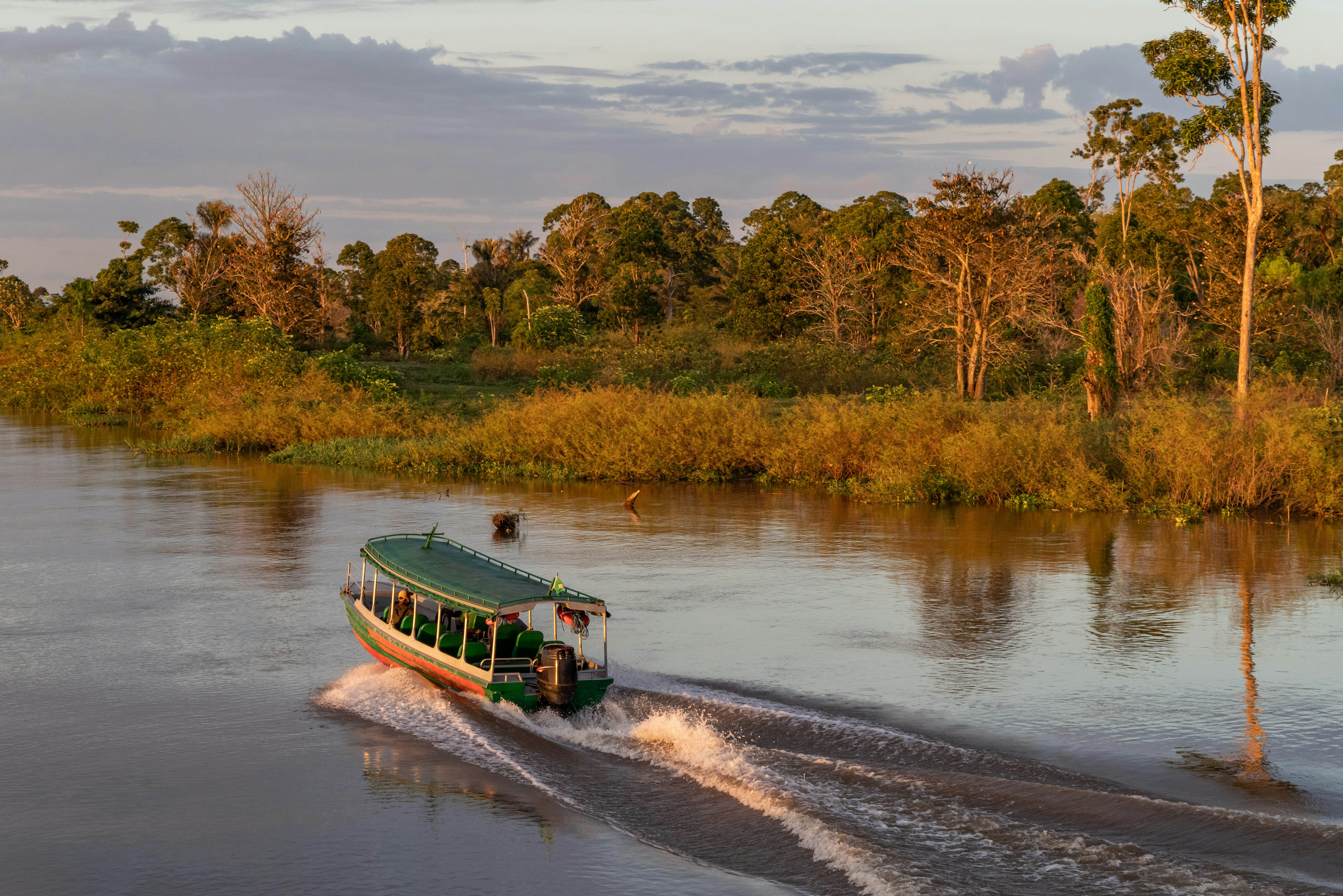 A ferry boat navigates a wide river at sunrise, trailing a wake in the still water. Trees and grasses bathed in golden light are visible on the riverbank