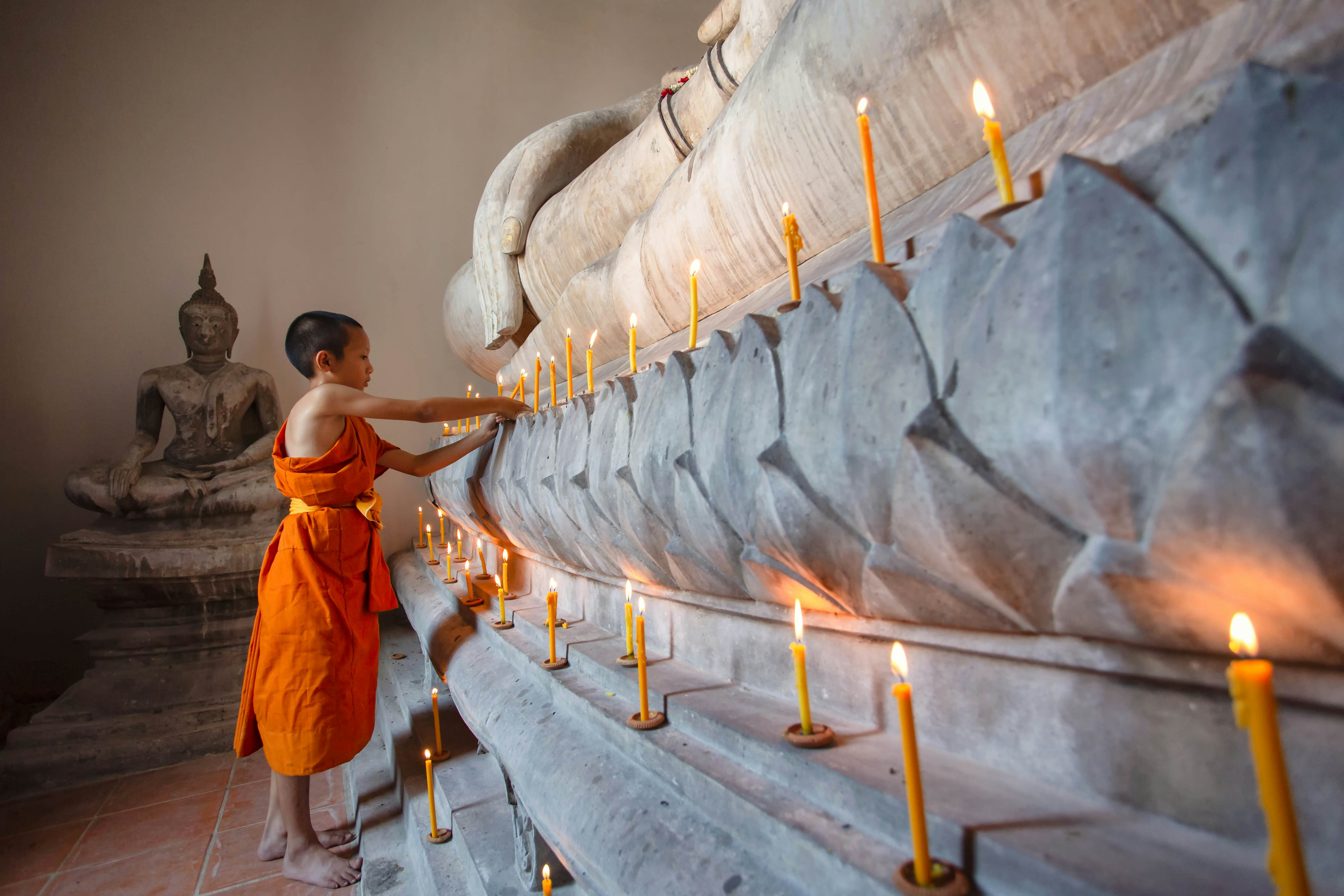 A young monk in a saffron robe lights a candle at the base of a Buddha statue in a temple.