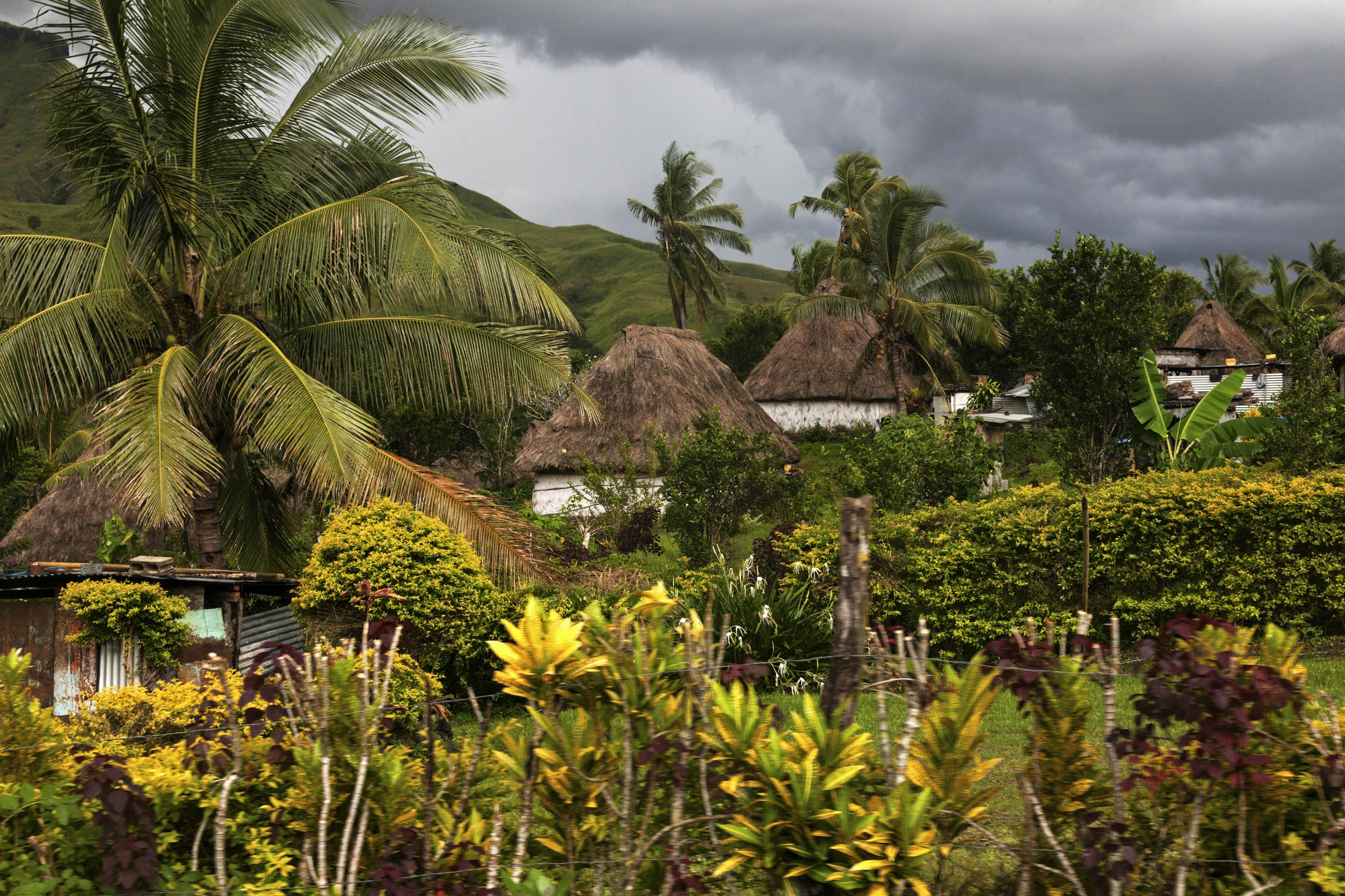 Thatched-roof buildings in a village in a mountainous tropical location filled with greenery. Storm clouds loom overhead.