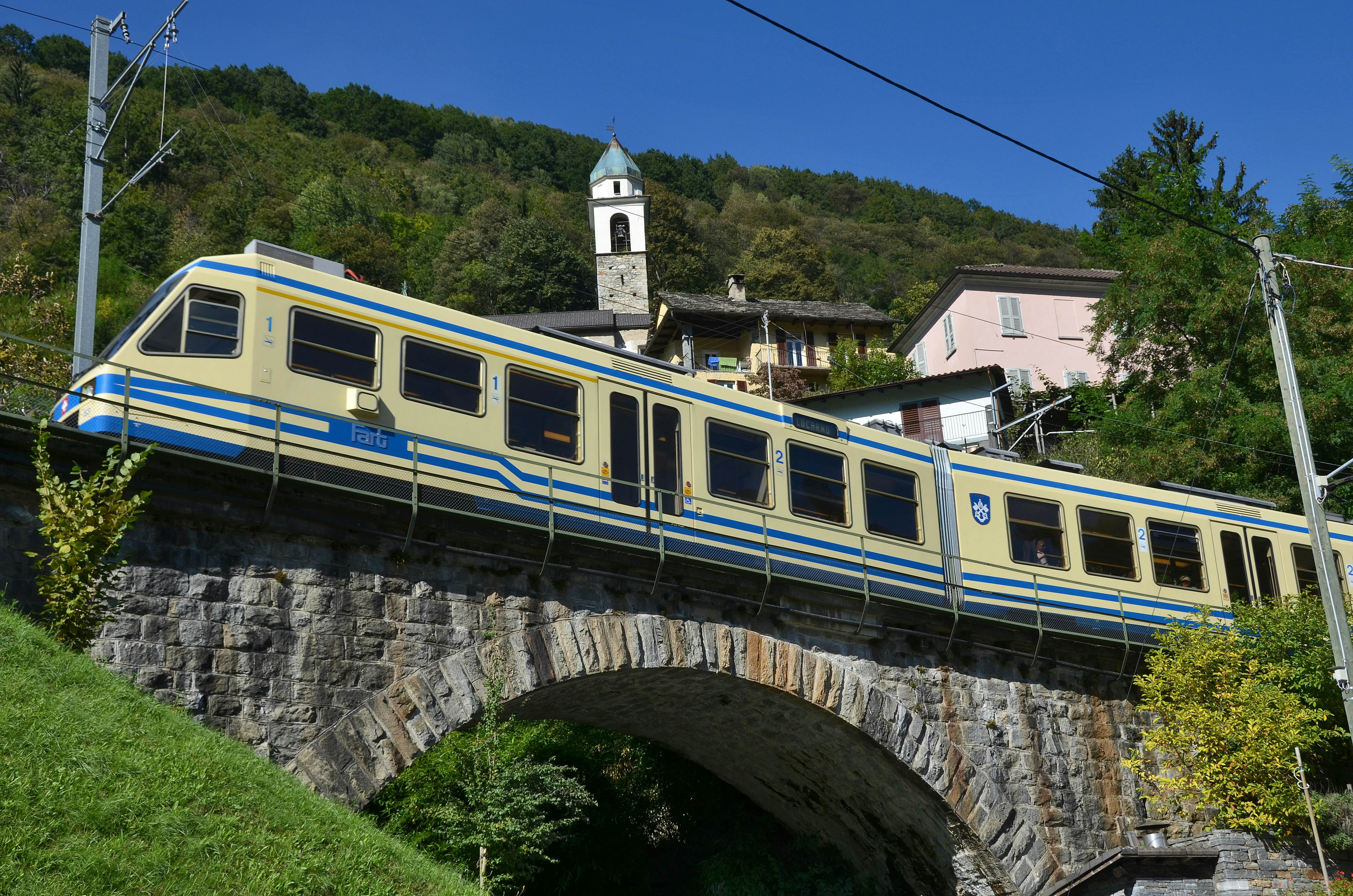 White and blue train crossing stone bridge with church spire and homes on a hill in the background with a forested hillside in the distance on a sunny day.