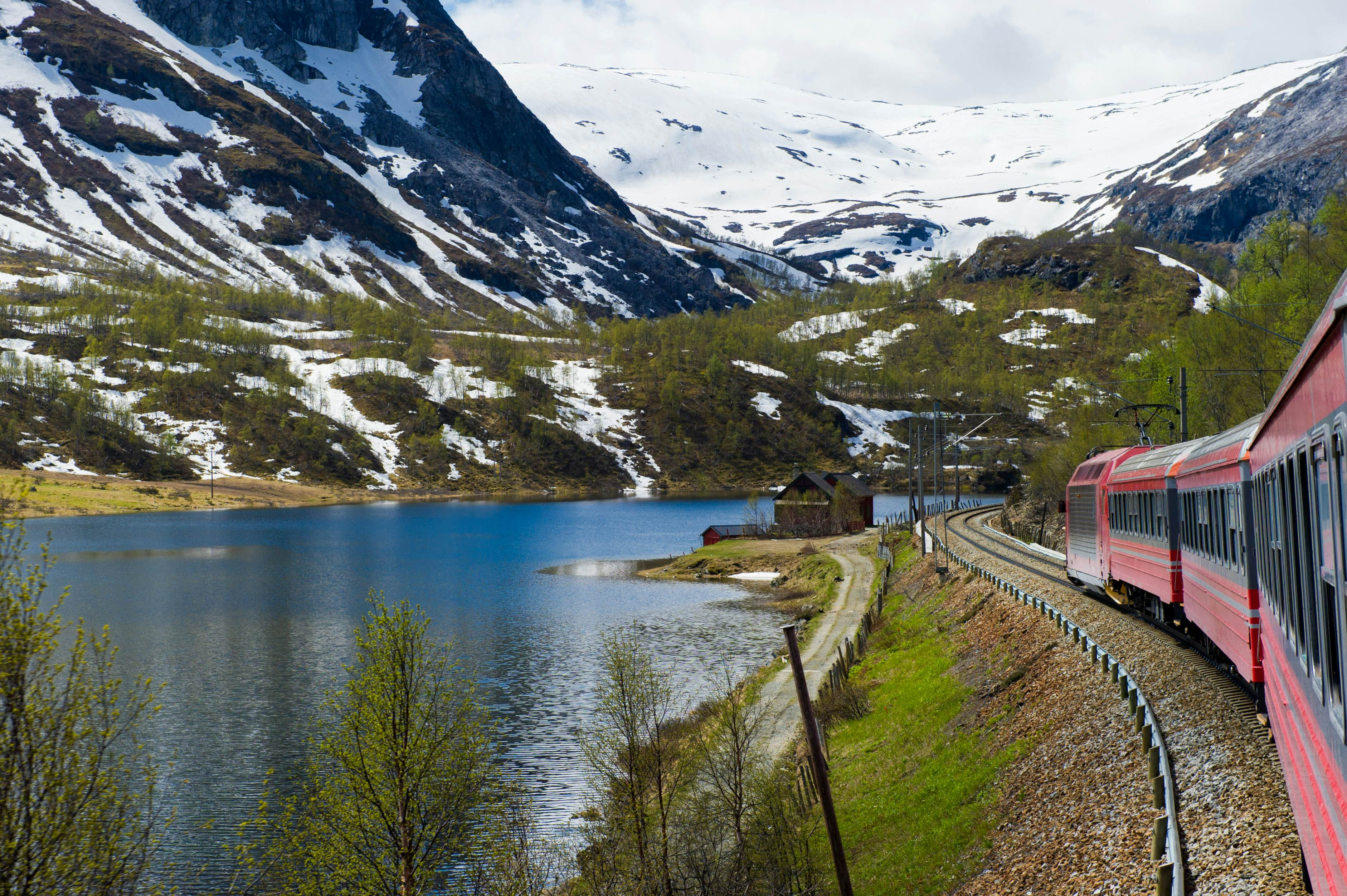 A red train passes snow-capped mountains and a river on a cloudy day.