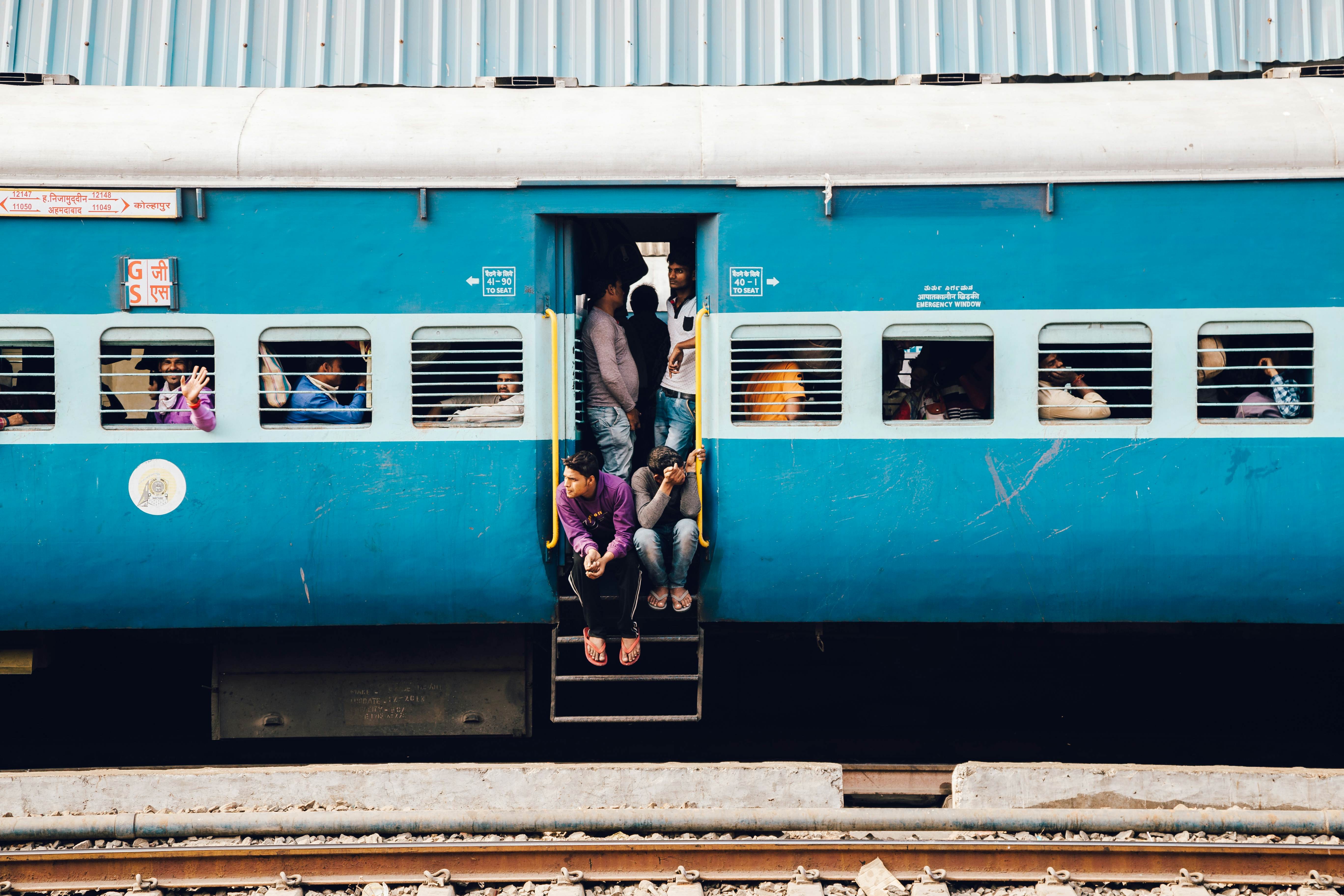 A stationary blue train in India, with passengers sitting and leaning out of the doorway and looking through the train