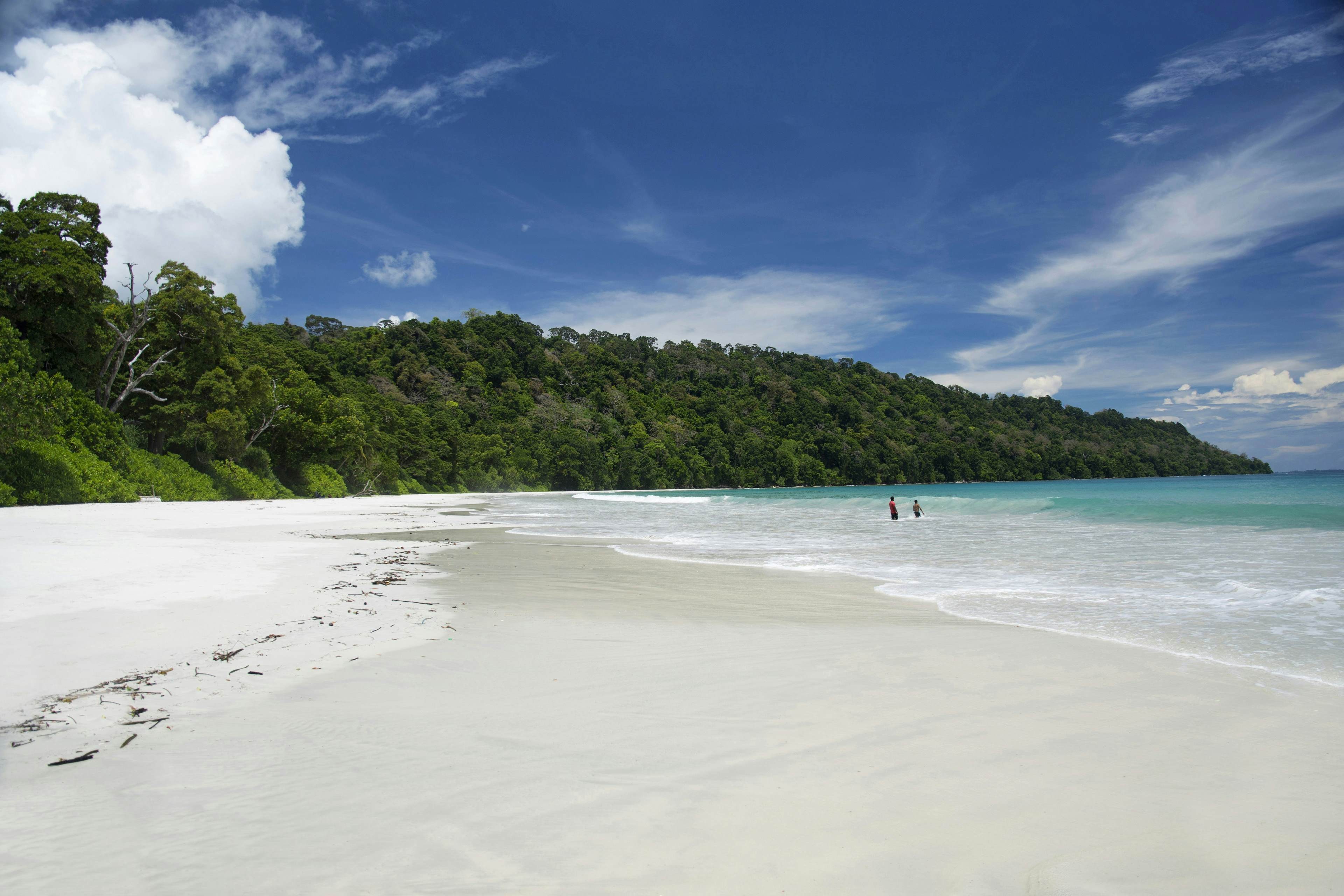 Two men kayak in the shallow waters of a serene beach on Havelock Island, part of the Andaman Islands. The beach features white sand and a forested backdrop.
