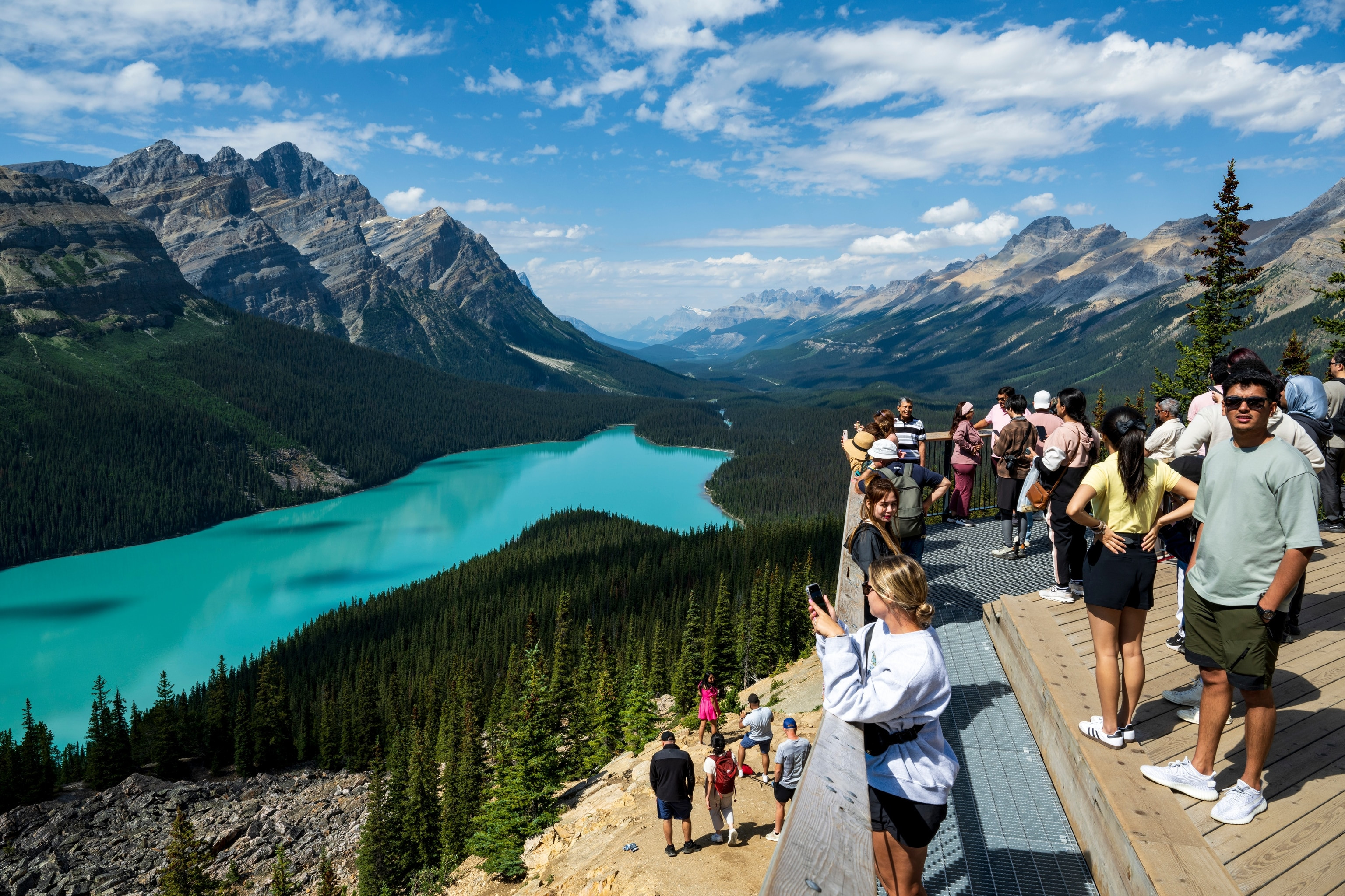 A group of tourists stands on a platform overlooking a bright blue lake surrounded by forest and mountains.