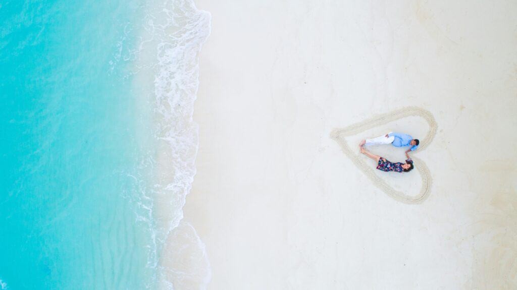 A couple walking on a beach with a sand heart in the foreground, capturing the romance and celebration of a honeymoon